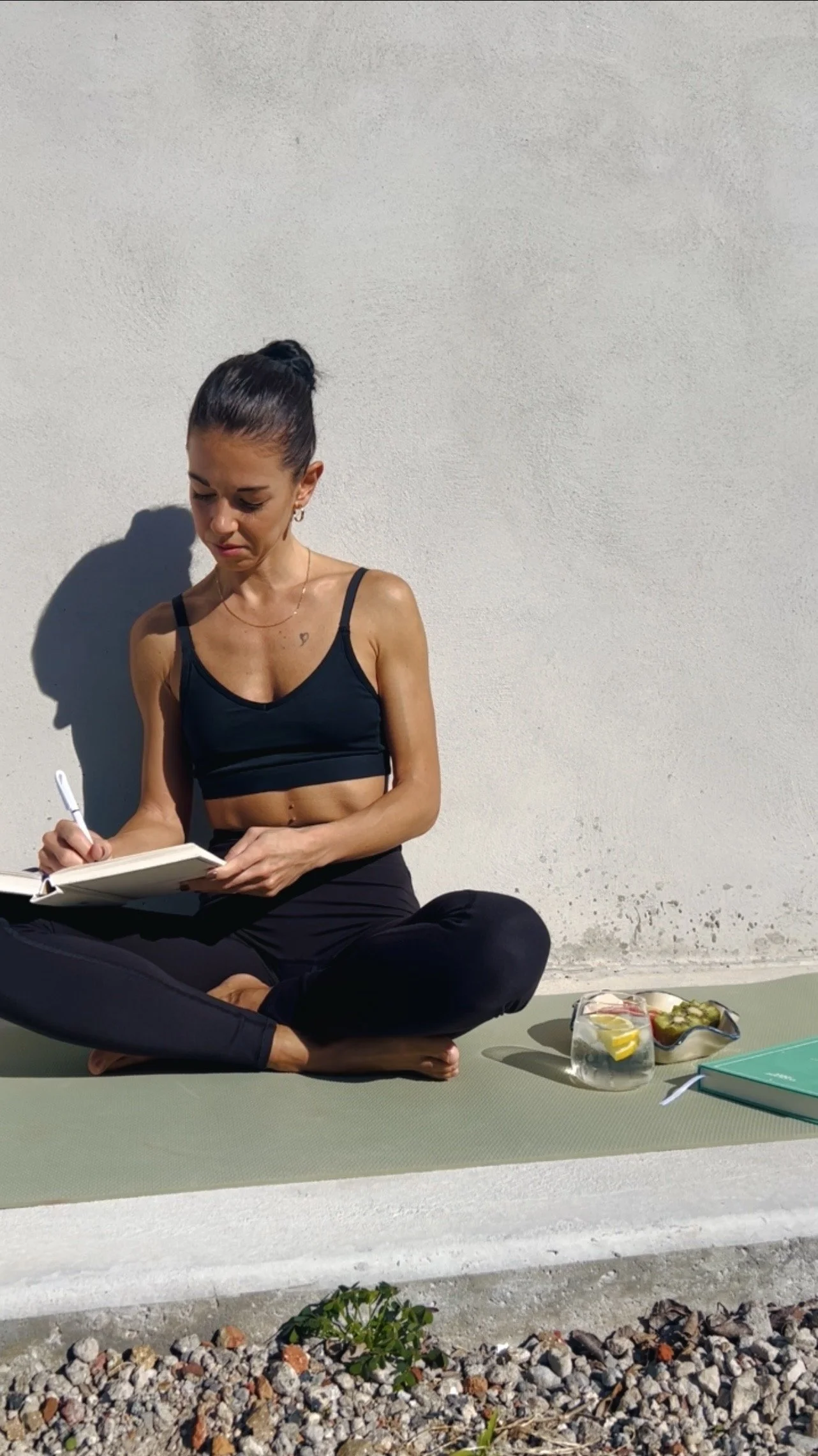 Woman in black workout clothes sitting cross-legged on a yoga mat, writing in a notebook outdoors against a plain white wall, with a glass container of water and snacks nearby. Journaling, sun, moments for yourself.