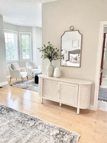 Bright living room with white armchairs, a window with blinds, a rug, and a hallway leading to a bedroom with a bed and window in the background.