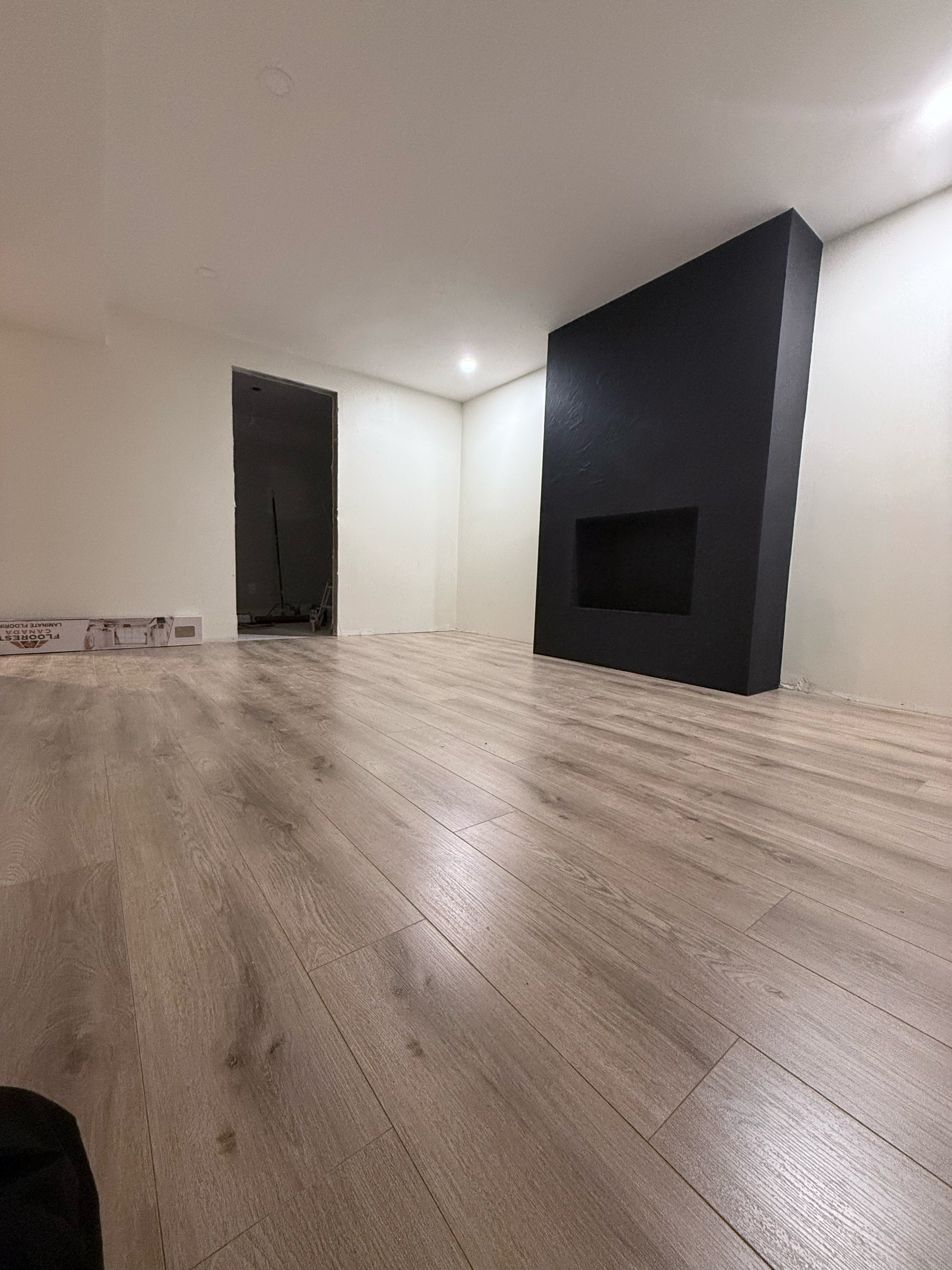 Unfinished living room with light wood flooring, white walls, a black accent wall with a built-in fireplace opening, and an open doorway leading to another room.