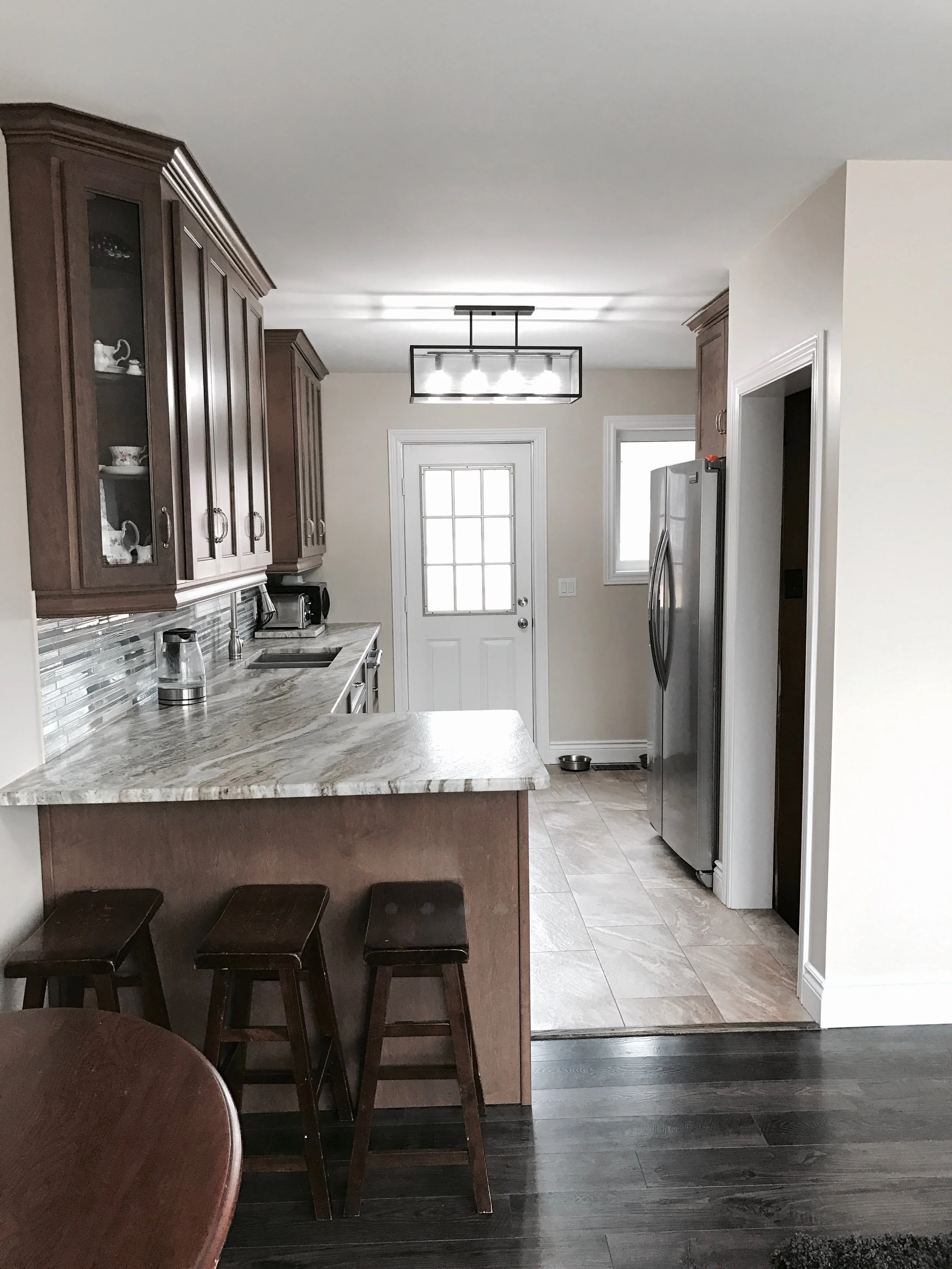 A kitchen with wooden cabinets, stainless steel appliances, a marble countertop, and three dark wooden barstools, with a door and window at the back.