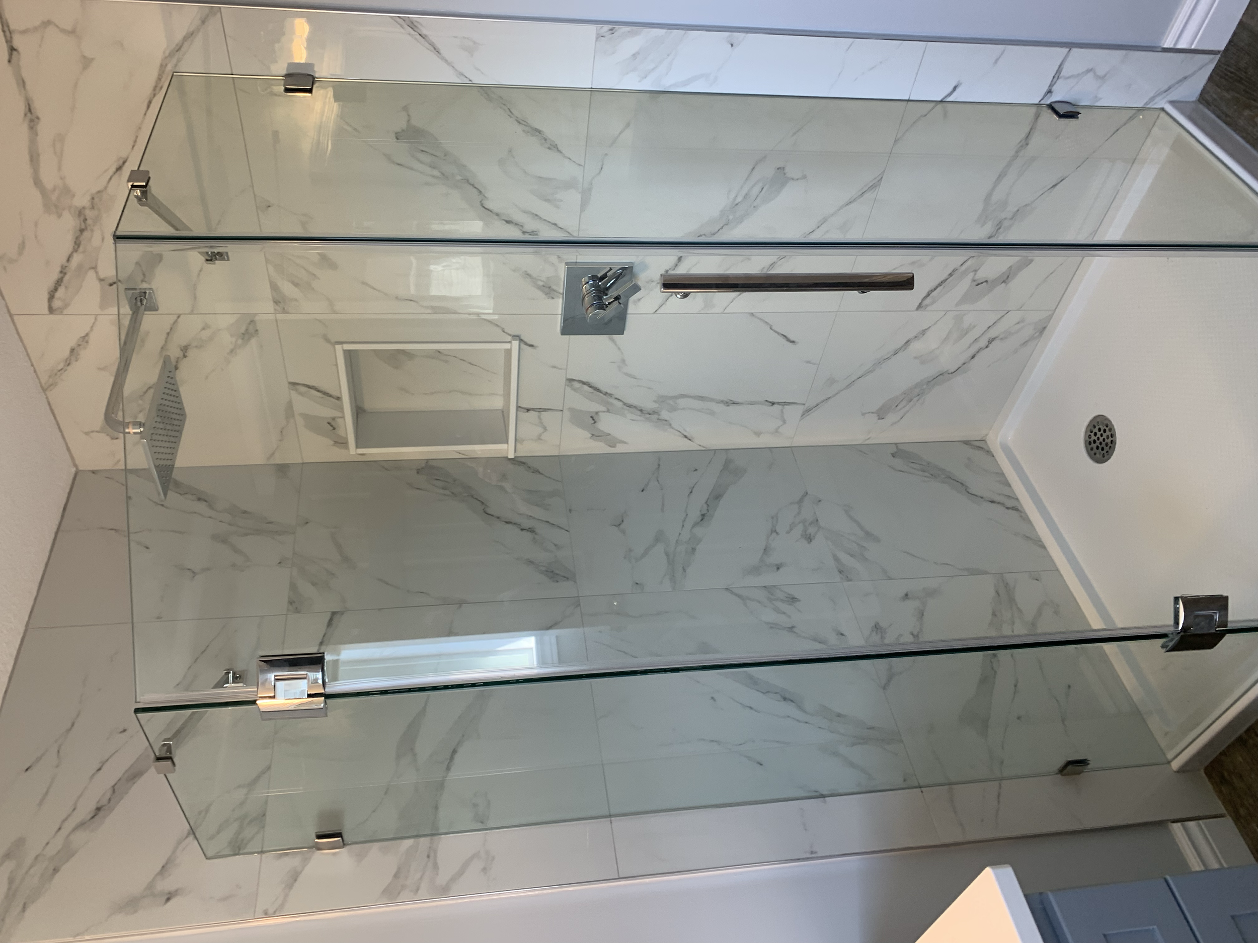 A modern walk-in shower with glass doors, white marble walls with gray veining, a rainfall showerhead, a hand-held shower wand, and a built-in shelf, with a drain on the floor.