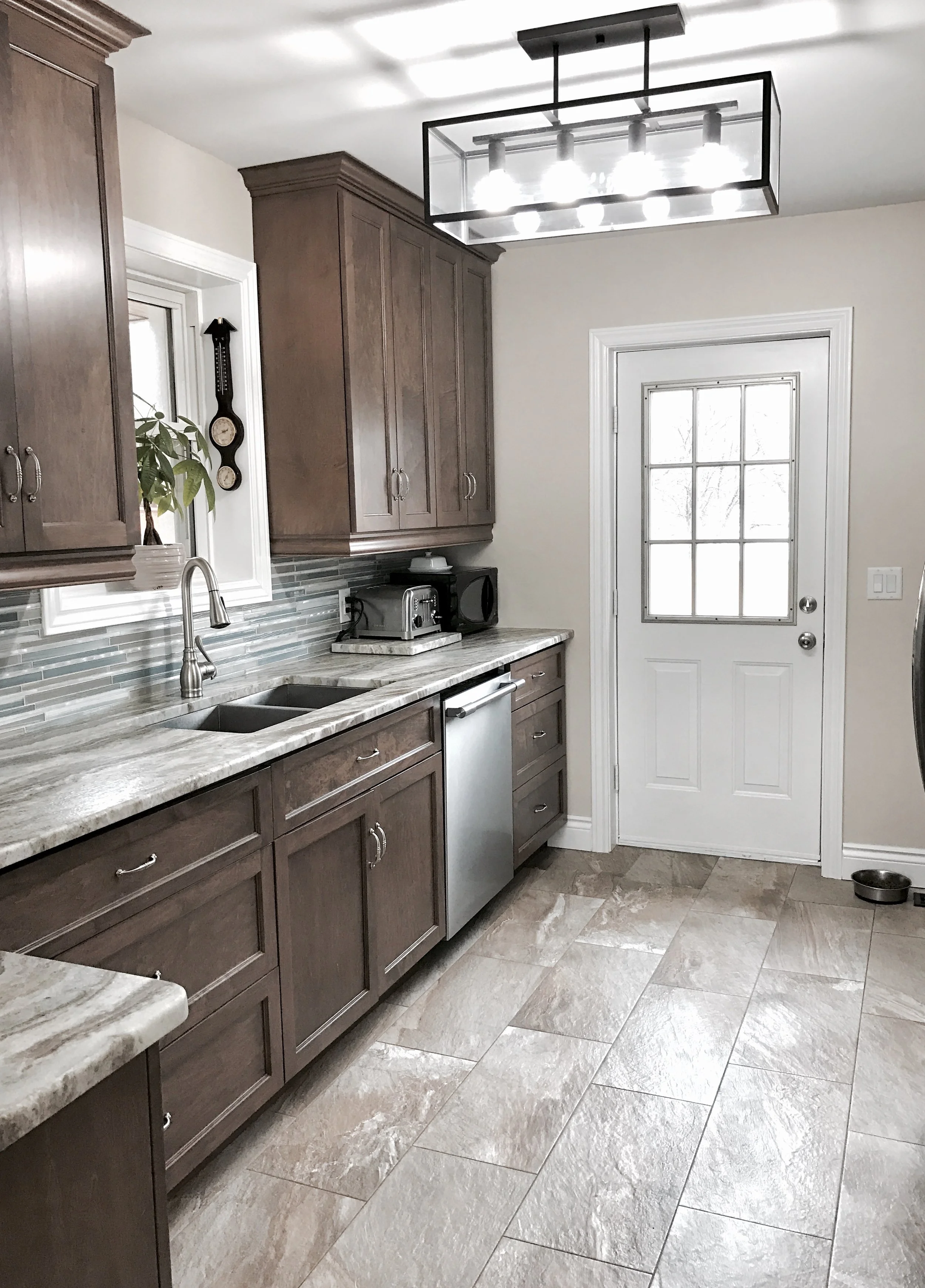 Kitchen with brown cabinets, granite countertops, tiled backsplash, and a white door with a window, modern light fixture on ceiling.