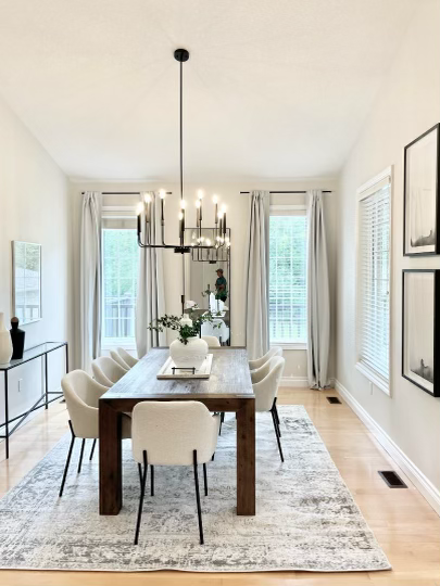 Dining room with a wooden table, beige chairs, black chandelier, large windows with white curtains, white walls with framed artwork, and a side table with decorative items.