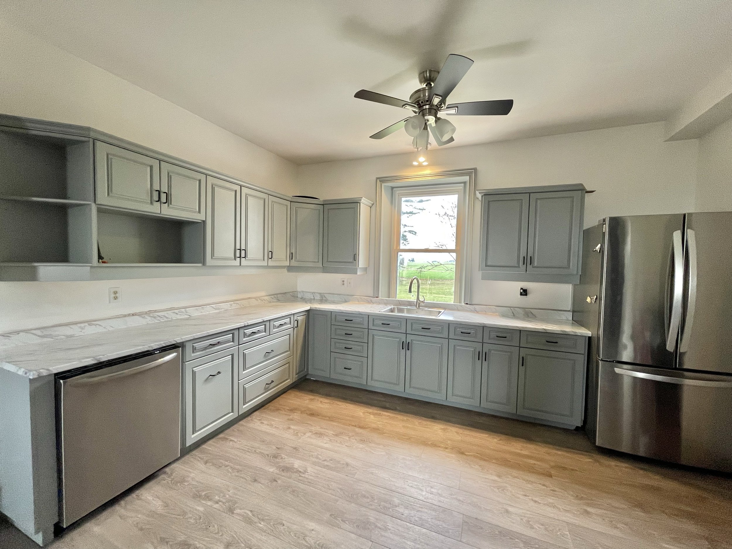 Modern kitchen with light gray cabinets, white marble countertops, stainless steel appliances including a fridge and dishwasher, a window over the sink, a ceiling fan, and wooden flooring.
