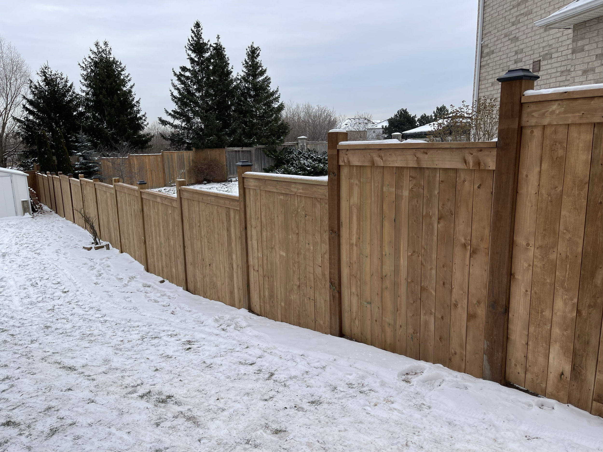 Wooden fence running along a snowy backyard with trees and a house in the background.
