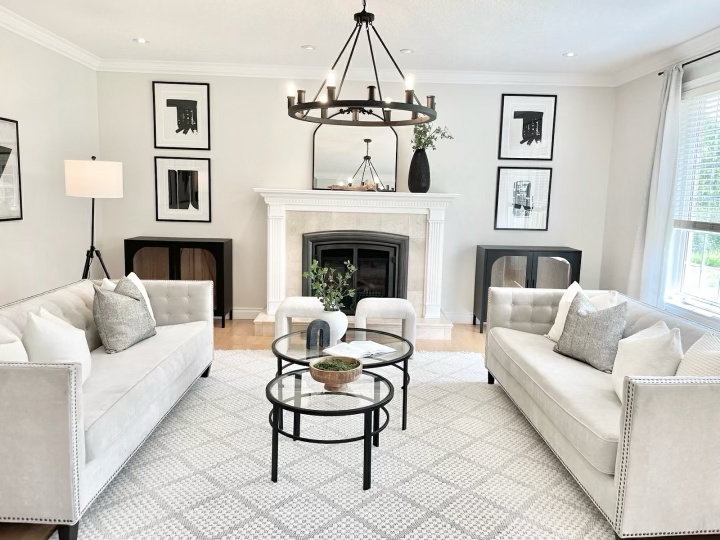 Bright living room with two white sofas, black and wood side tables, a fireplace with a mirror and decor on the mantel, black and white wall art, a large window with white curtains, and a black chandelier overhead.