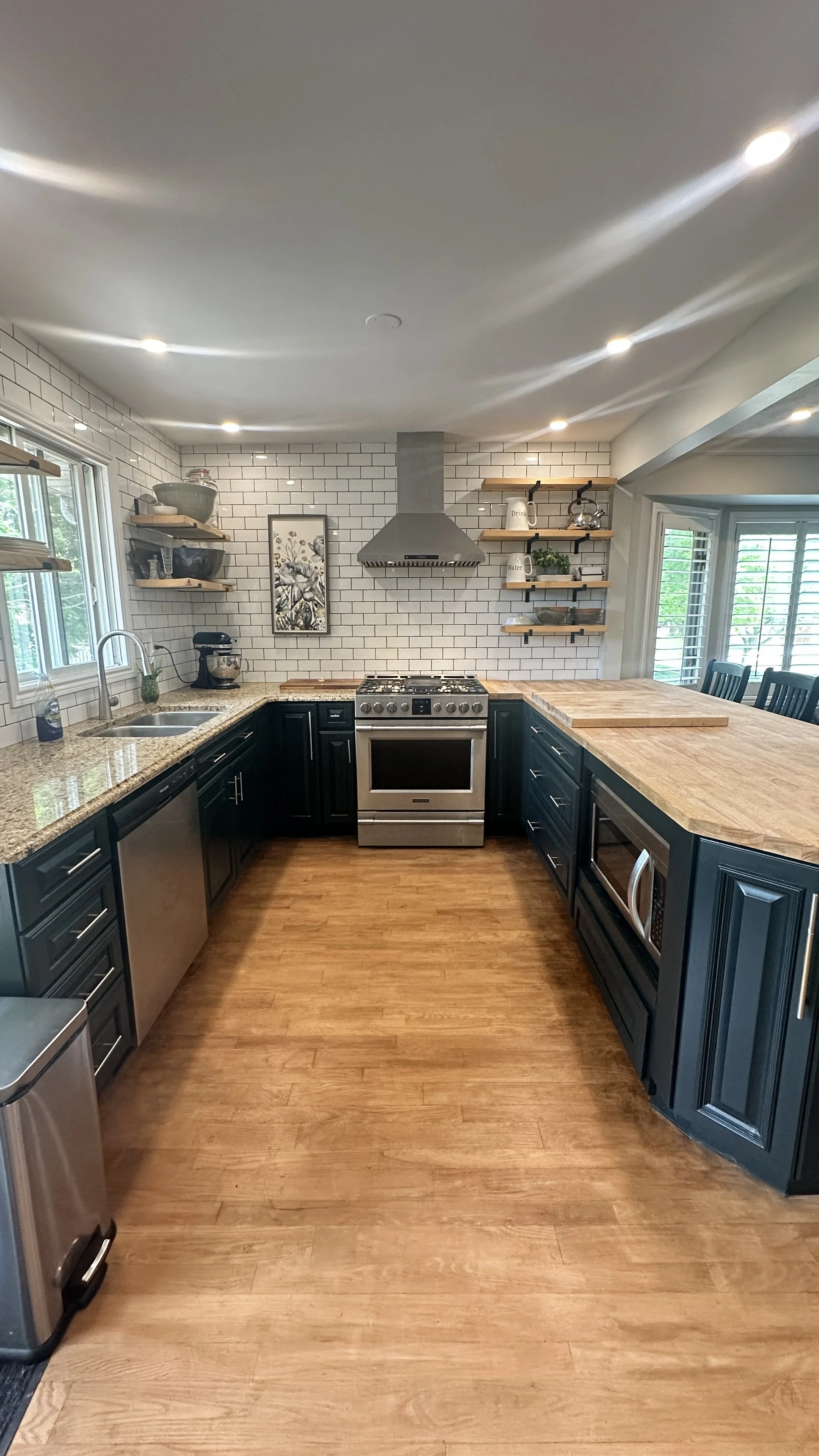 Modern kitchen with black cabinets, wooden countertops, white subway tile backsplash, stainless steel appliances, open wooden shelves, and hardwood floors.