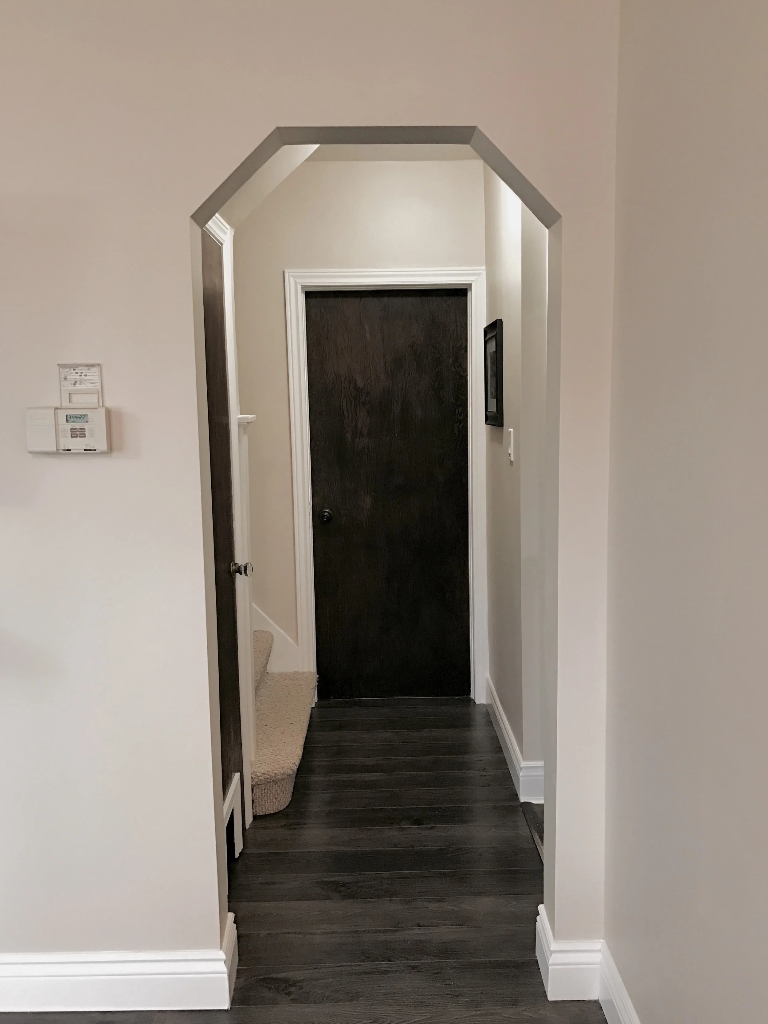 A hallway with dark wood flooring, leading to a closed dark brown door, framed by white molding. The hallway features an archway with a geometric shape, beige walls, and a small section of carpeted stairs on the left. There is a thermostat on the lef