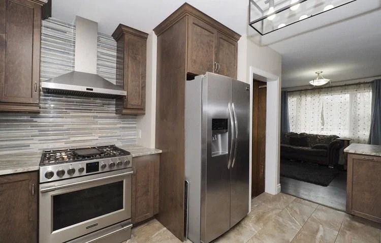 Kitchen with stainless steel refrigerator, gas stove, wooden cabinets, tile backsplash, and a view into a living room with curtains and a sofa.