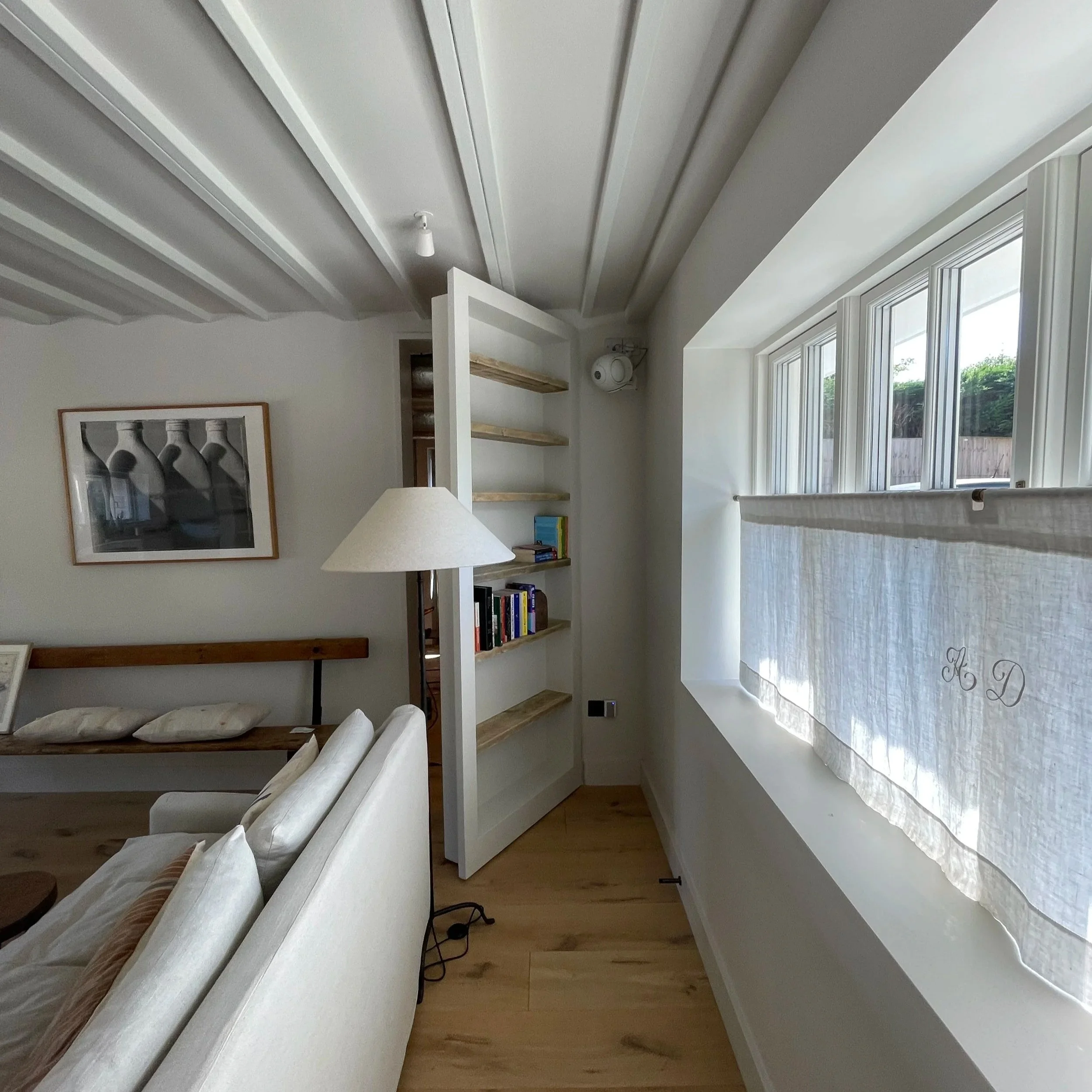 A cozy living room corner featuring a white sofa, a wall-mounted picture, a white open bookshelf with books, and a window with a light-colored curtain in a bright, modern style.