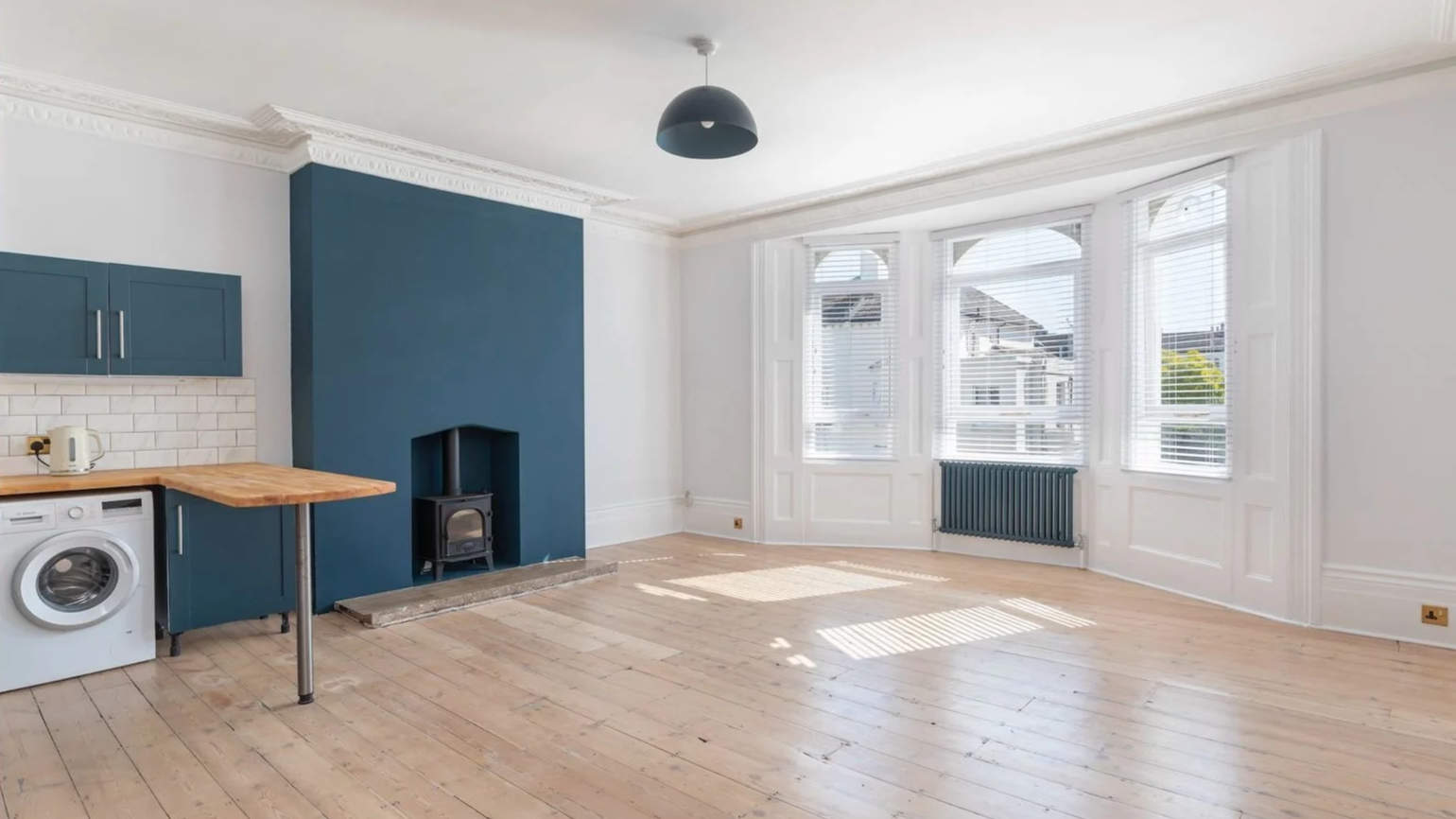 Empty living room with large bay windows, white walls, light wooden floors, blue accent wall, small fireplace, washing machine, small table, kettle, and black ceiling light.