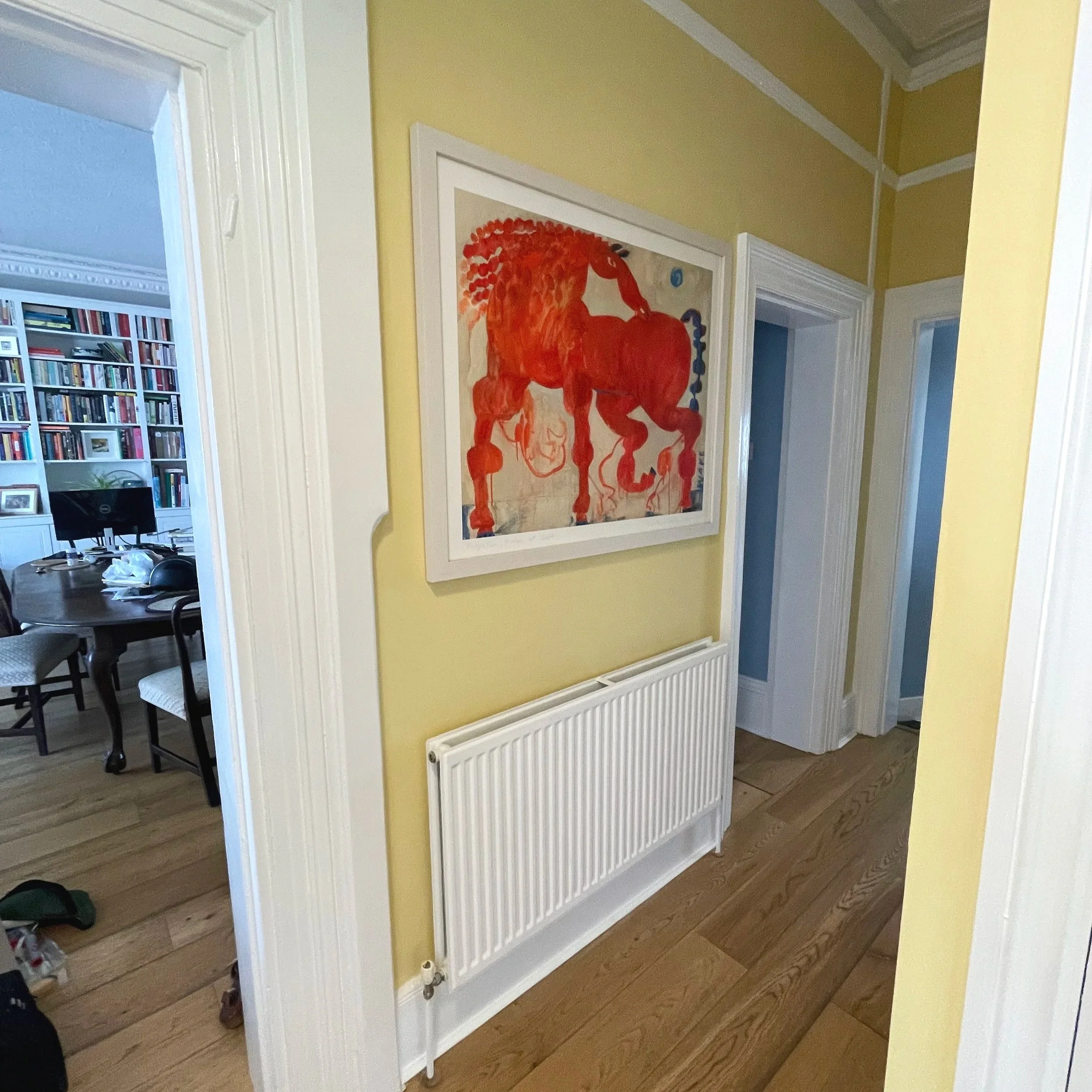 A hallway with yellow walls, a white radiator, and a framed painting of a red horse. A doorway to a room with bookshelves and a computer is visible on the left.