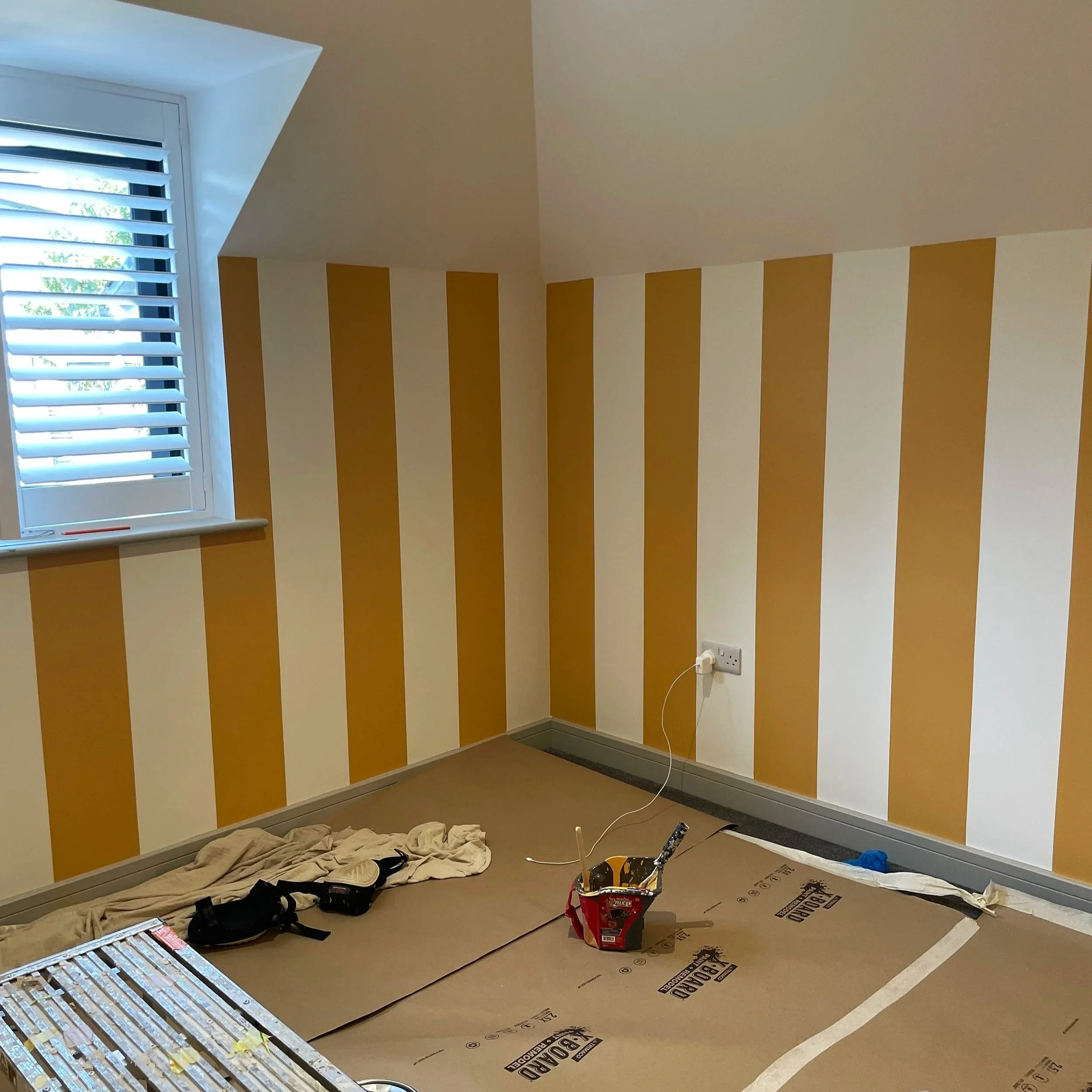 A room with a window on the left, yellow and white vertical striped walls, and construction tools on the floor, indicating ongoing decorating or renovation work.