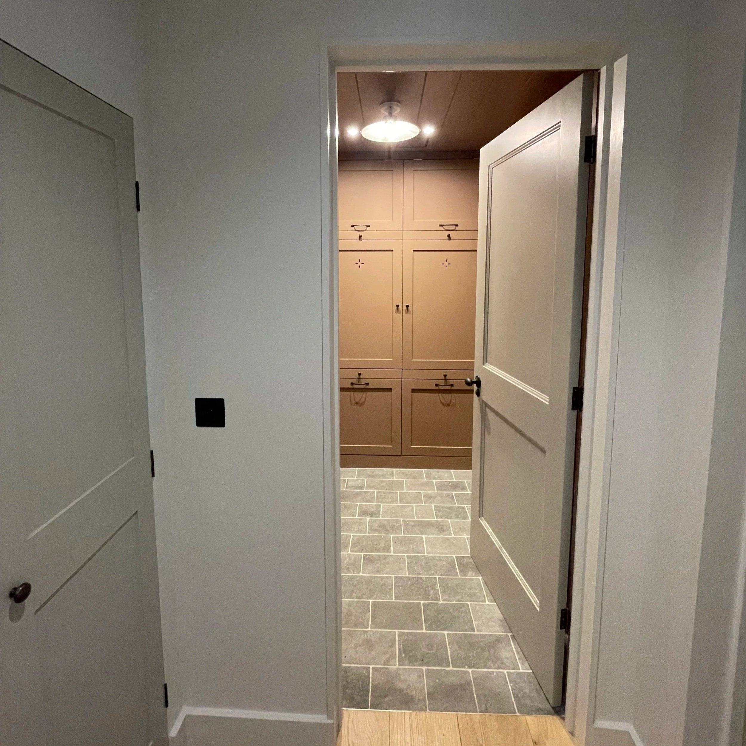 View of a door opening to a room with grey tile flooring and beige built-in cabinets with black handles on the back wall.