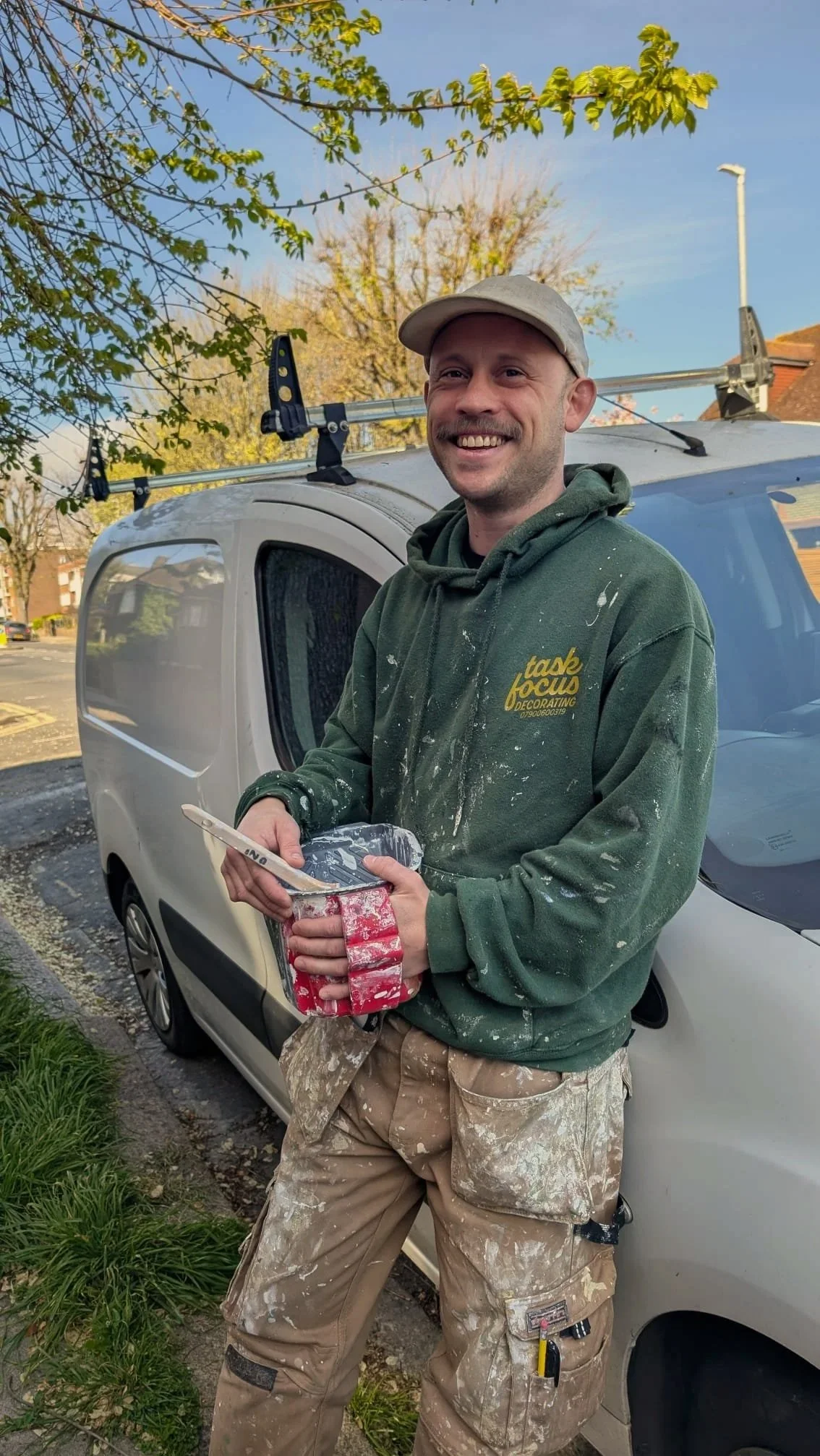A man smiling and holding a bowl of paint with a paintbrush in front of a white work van, with paint splatters on his green hoodie and beige work pants, standing outdoors on a street with trees and houses.