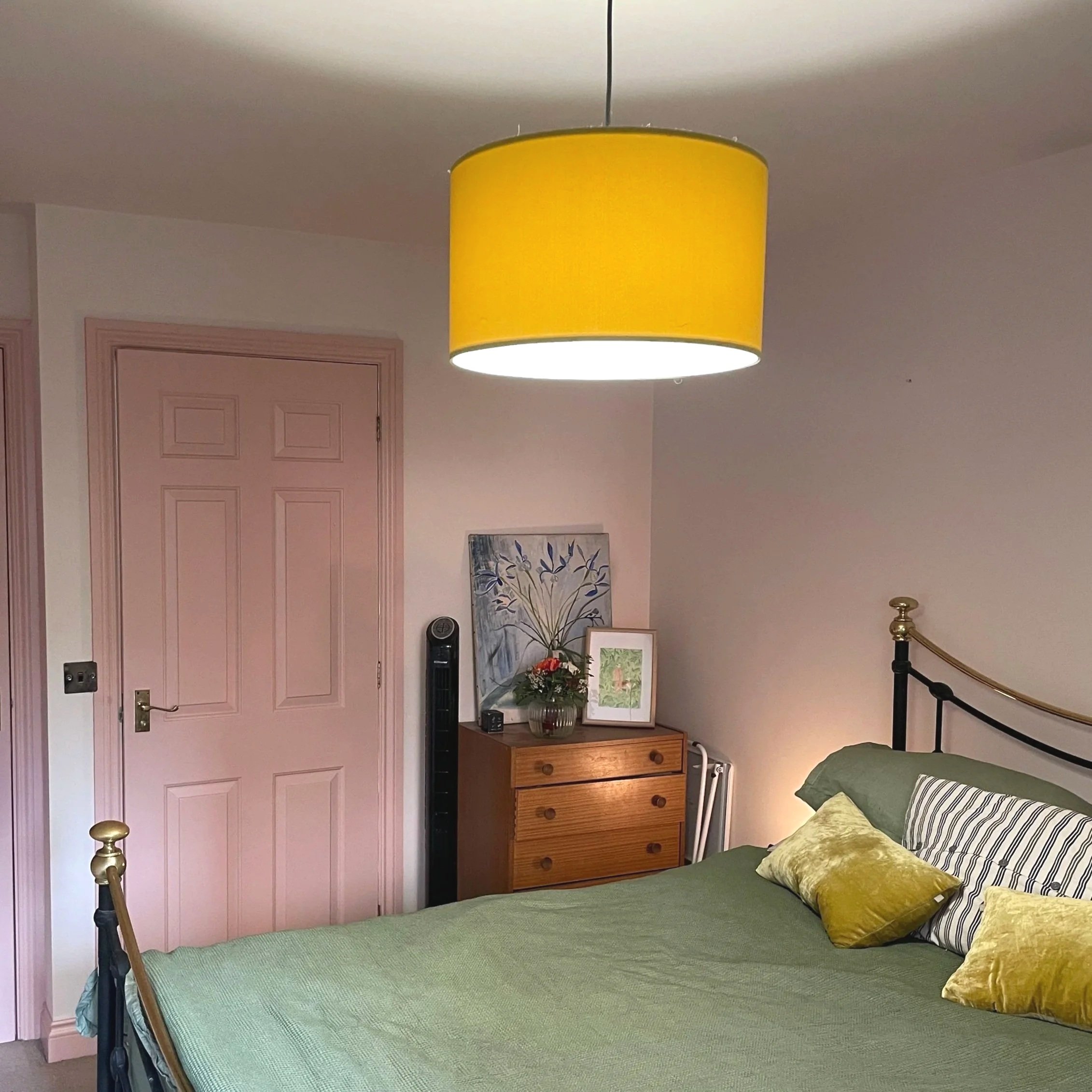 A bedroom with a green bedspread, decorative pillows, a black metal bed frame with gold accents, a wooden dresser with artwork and a flower vase, a pink door, and a large yellow ceiling lampshade.