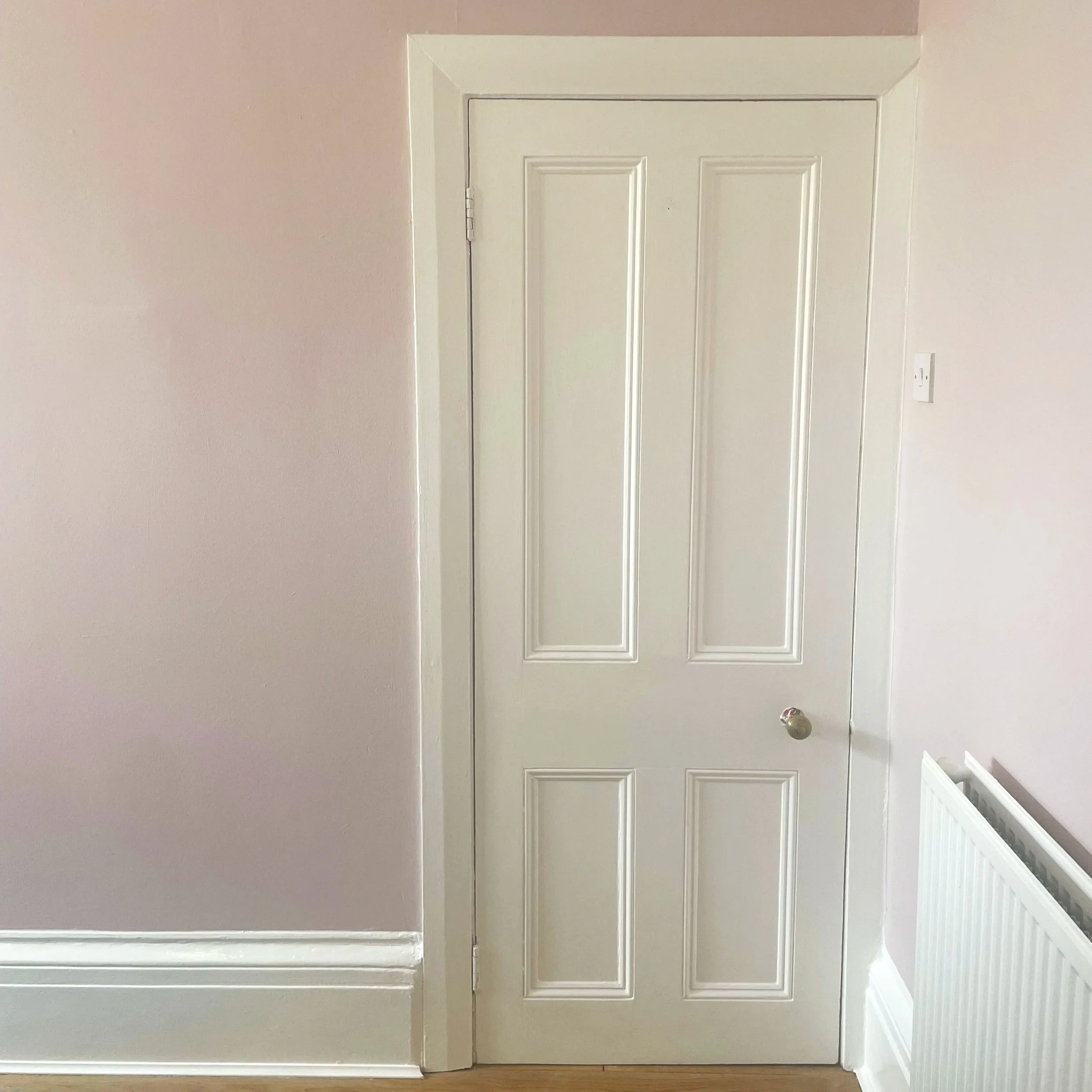 Closed white interior door with four raised panels, a silver doorknob, white trim, pink wall, beige baseboard, and part of a white radiator on the right.