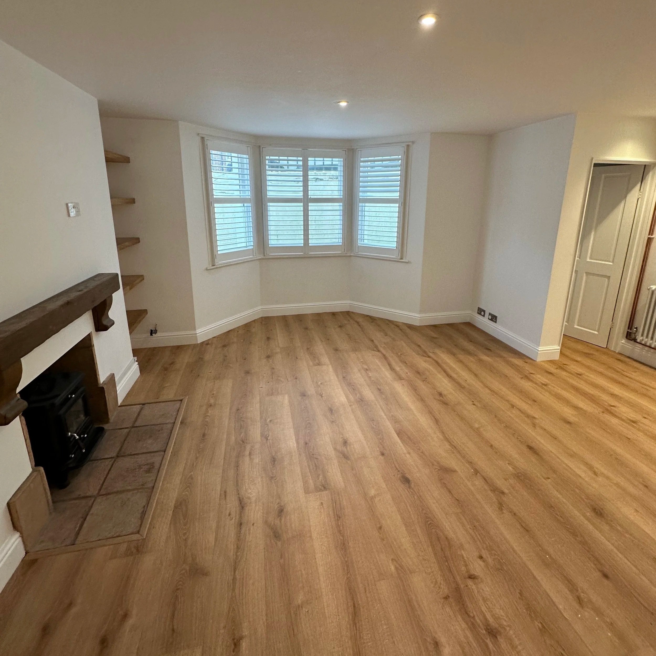 An empty living room with wood flooring, white walls, and a large bay window with blinds. There are built-in shelves on the left and a small fireplace with a tiled hearth. Recessed ceiling lights and a radiator are visible.