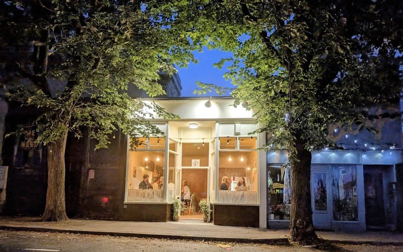 Street view of a busy restaurant with large front windows, illuminated from inside, at night. Two trees frame the restaurant, with string lights hanging outside, and people are dining inside.