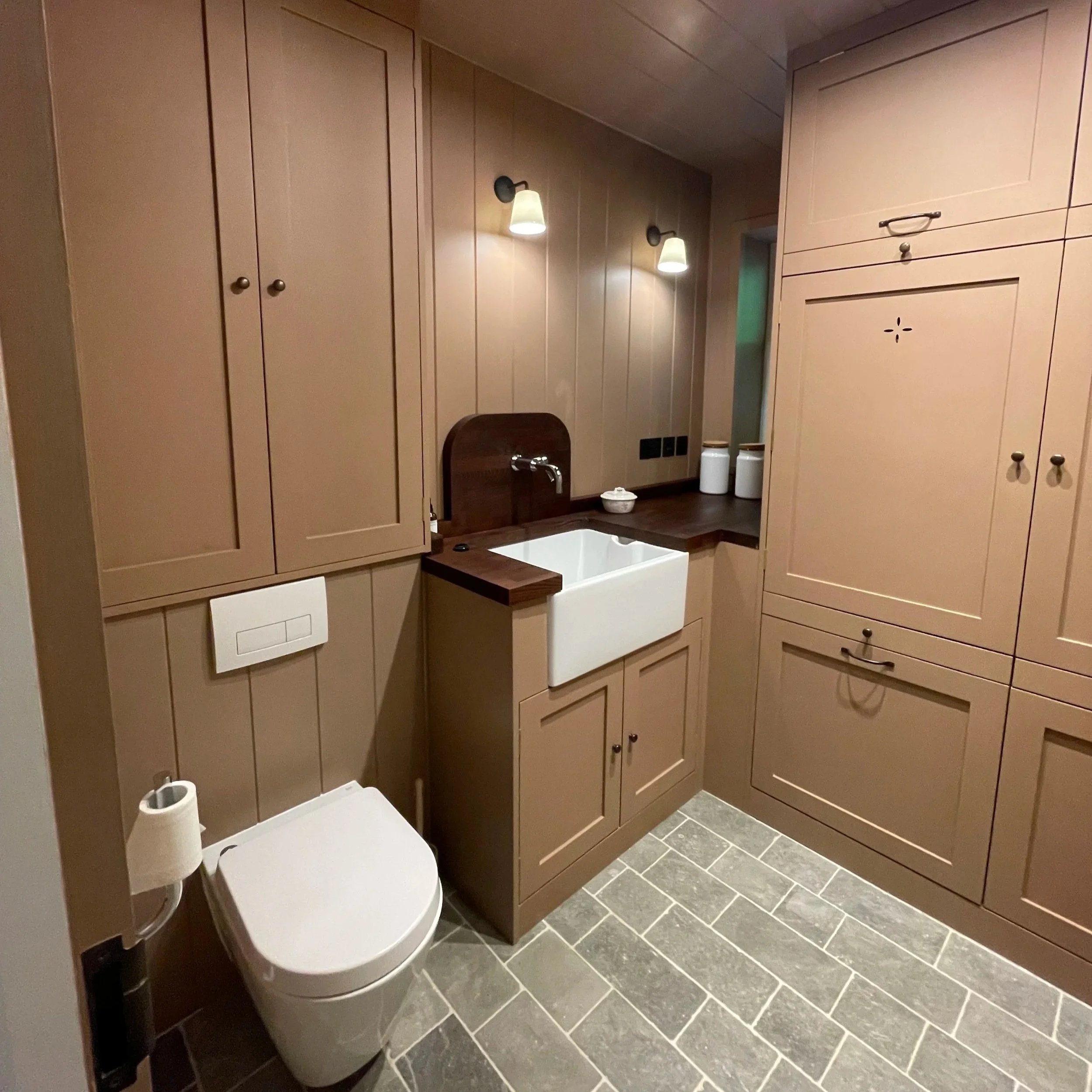 A modern bathroom featuring tan cabinetry, a white utility sink with a wooden countertop, wall-mounted lighting, and a toilet with a wall-mounted flush panel. The floor has gray stone tiles, and there are two white canisters and a small bowl on the countertop.