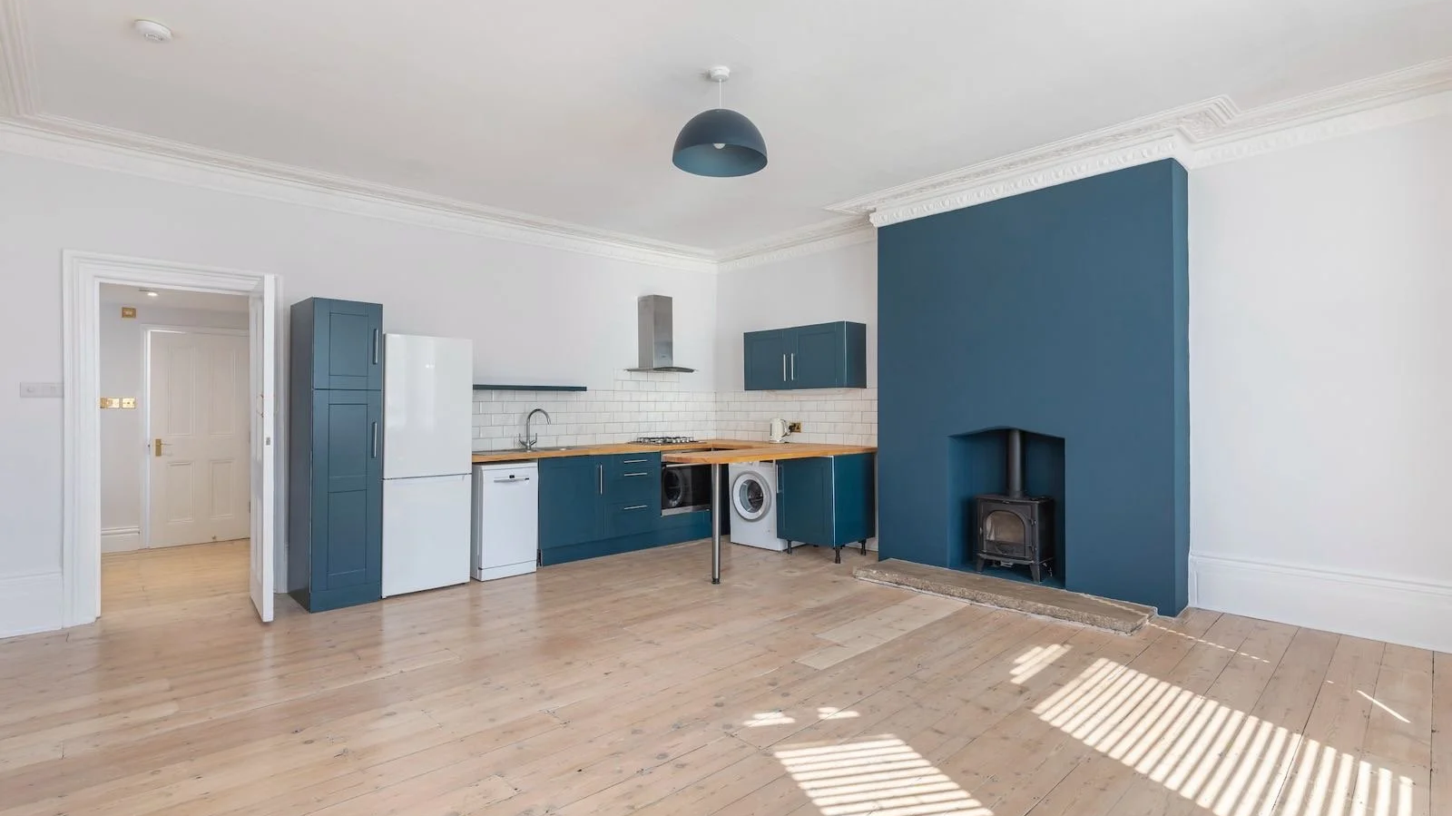 Open-plan kitchen with blue cabinets, white subway tile backsplash, wooden countertops, a white refrigerator, washing machine, and a stove, with a fireplace in the corner and light wood flooring.
