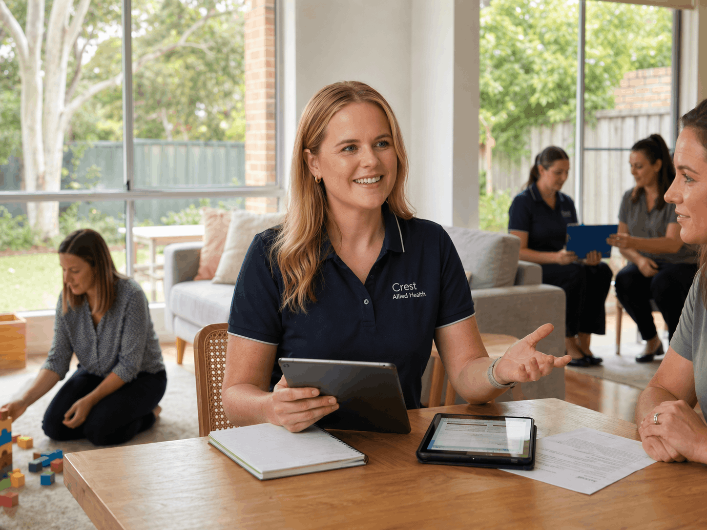 A woman wearing a navy Blue polo with 'Crest Allied Health' embroidered on it is talking to another person at a table in a bright living room with large windows. Other women are in the background, some sitting on the floor and some sitting on chairs, engaged in various activities.