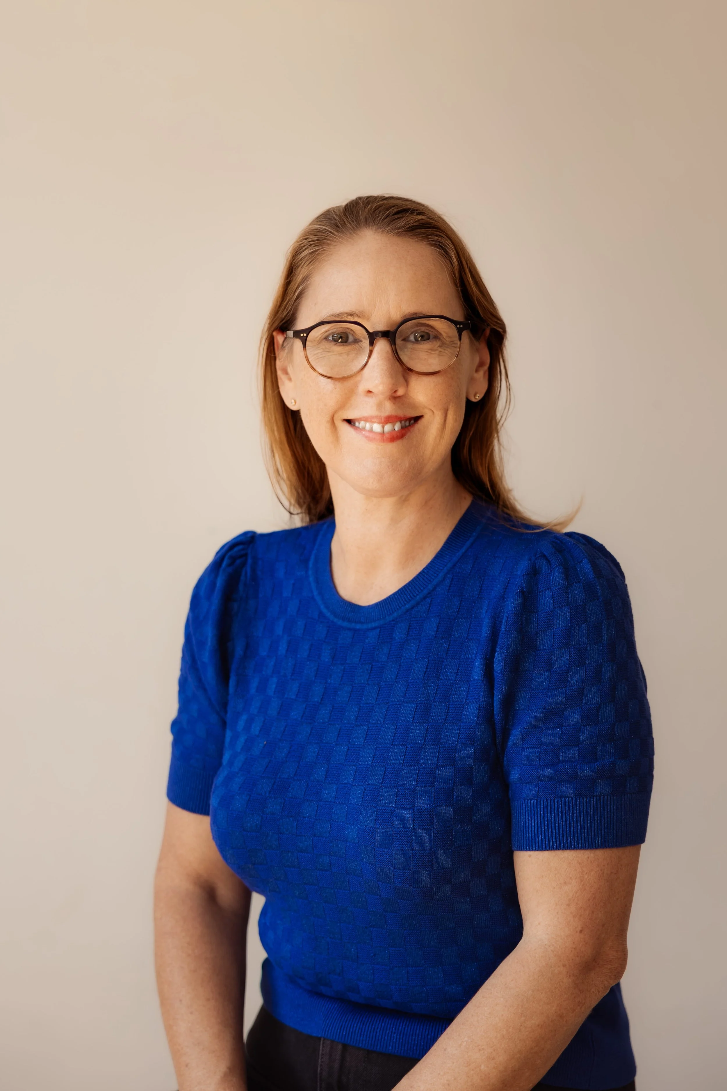 A woman with red hair, glasses, and a blue textured short-sleeve top smiling at the camera against a plain neutral background.