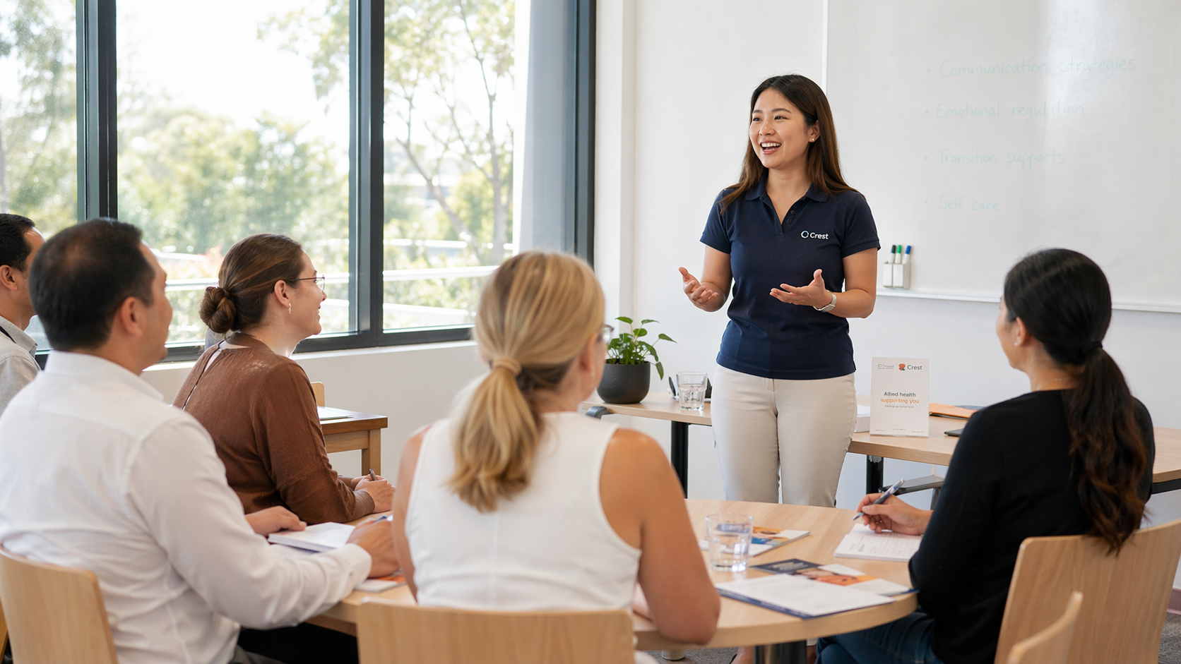 A woman stands at the front of a classroom giving a presentation to five seated adults, with a whiteboard and large windows behind her.