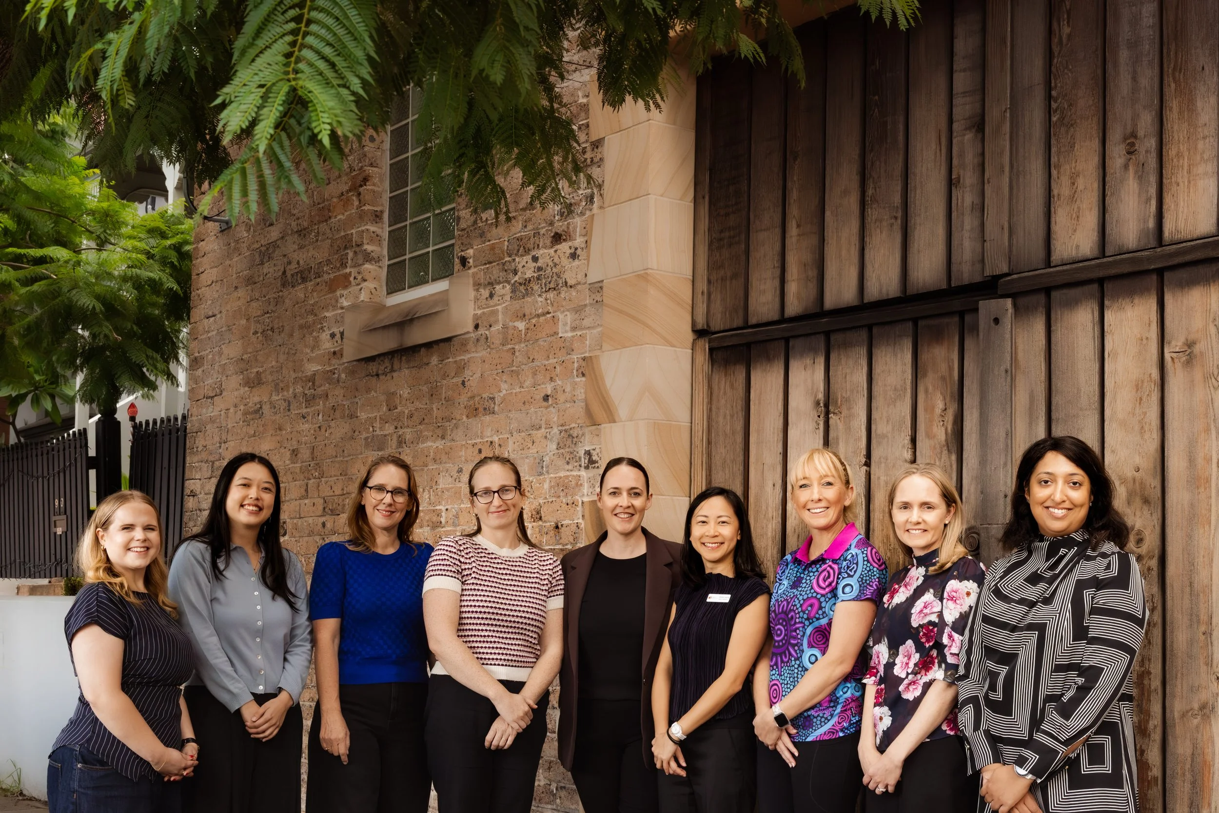 Group of diverse women standing outdoors in front of a brick and wood wall, smiling at the camera.