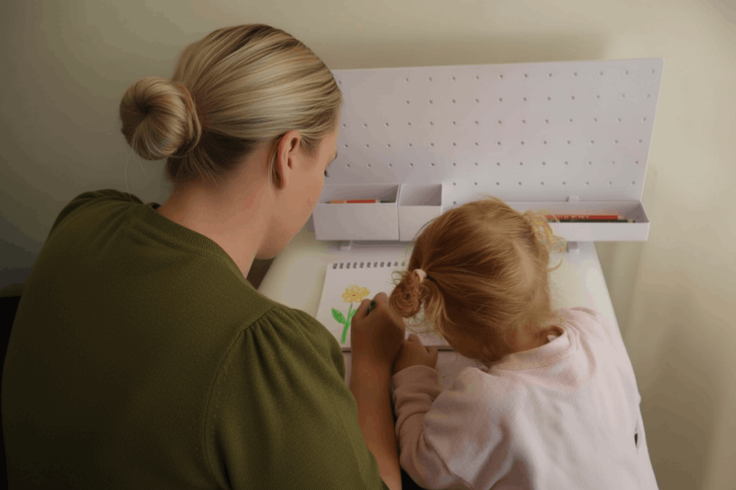 A woman with blonde hair tied in a bun leaning over a young girl with red hair drawing a flower on paper.