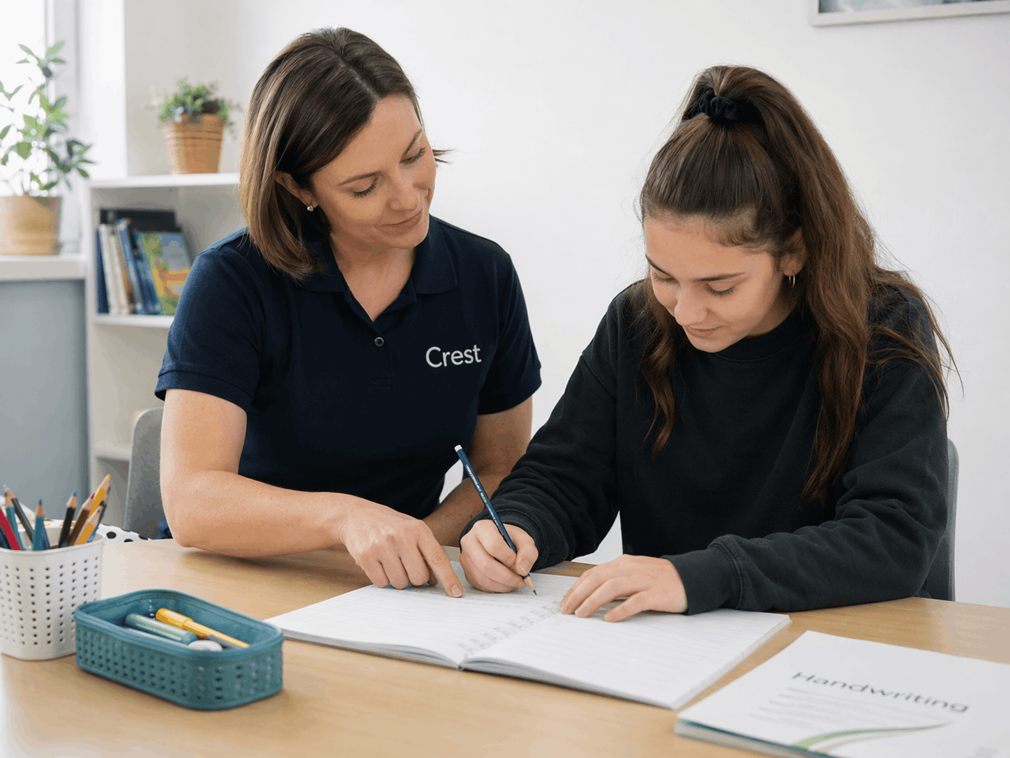A woman in a navy polo shirt with the word 'Crest' on it is helping a young girl with her handwriting. They are seated at a desk with a notebook, stationery, and a book titled 'Handwriting.'
