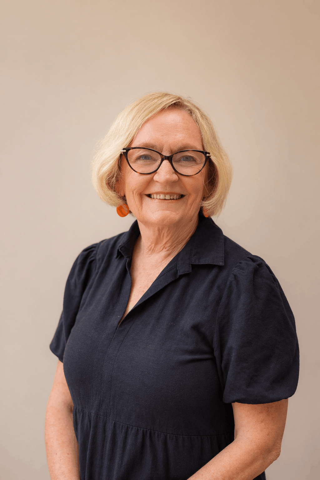 A smiling middle-aged woman with blonde hair, glasses, and orange earrings, wearing a navy blue dress, standing against a plain beige background.