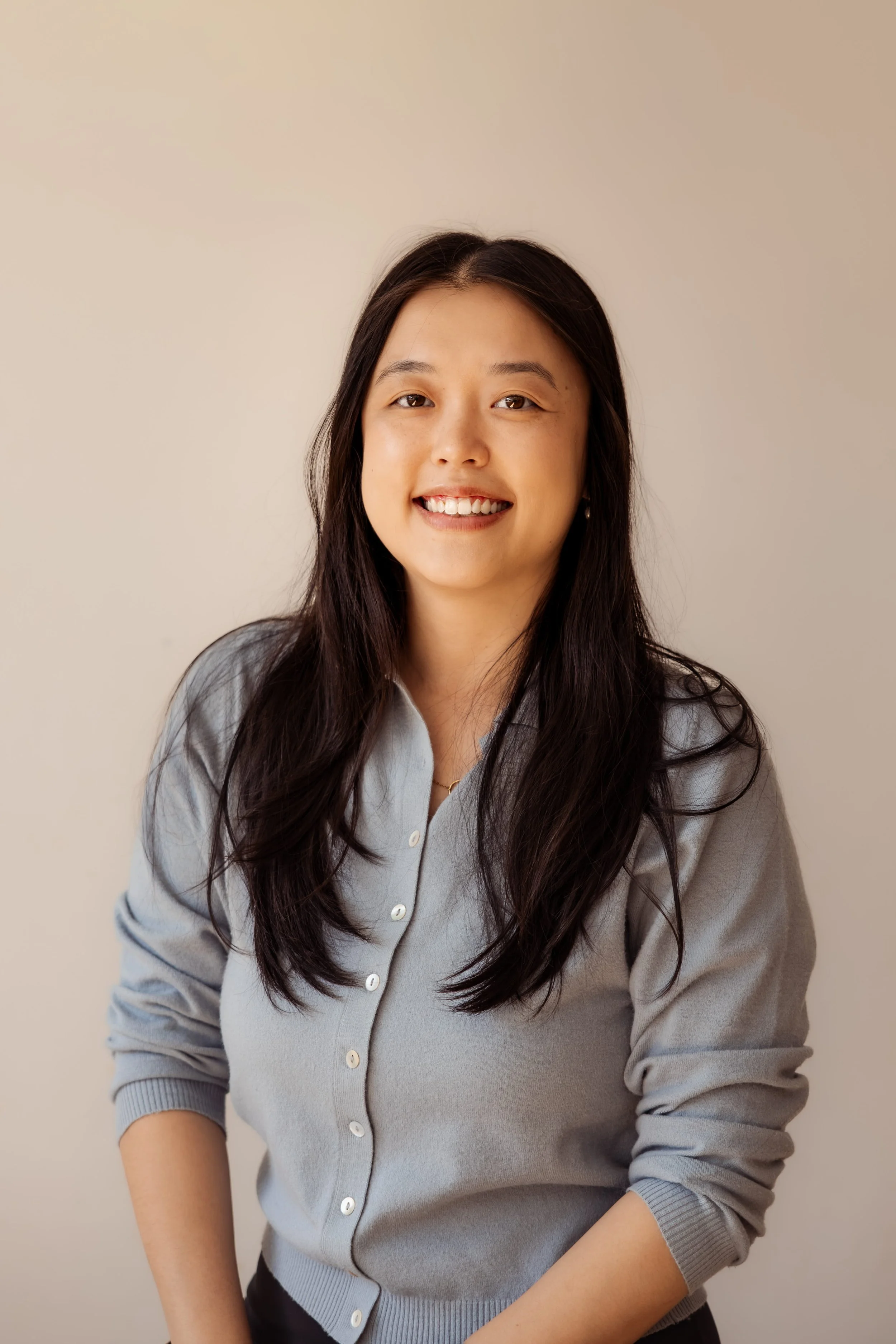 A woman with long dark hair wearing a light gray button-up sweater, smiling, against a plain neutral background.