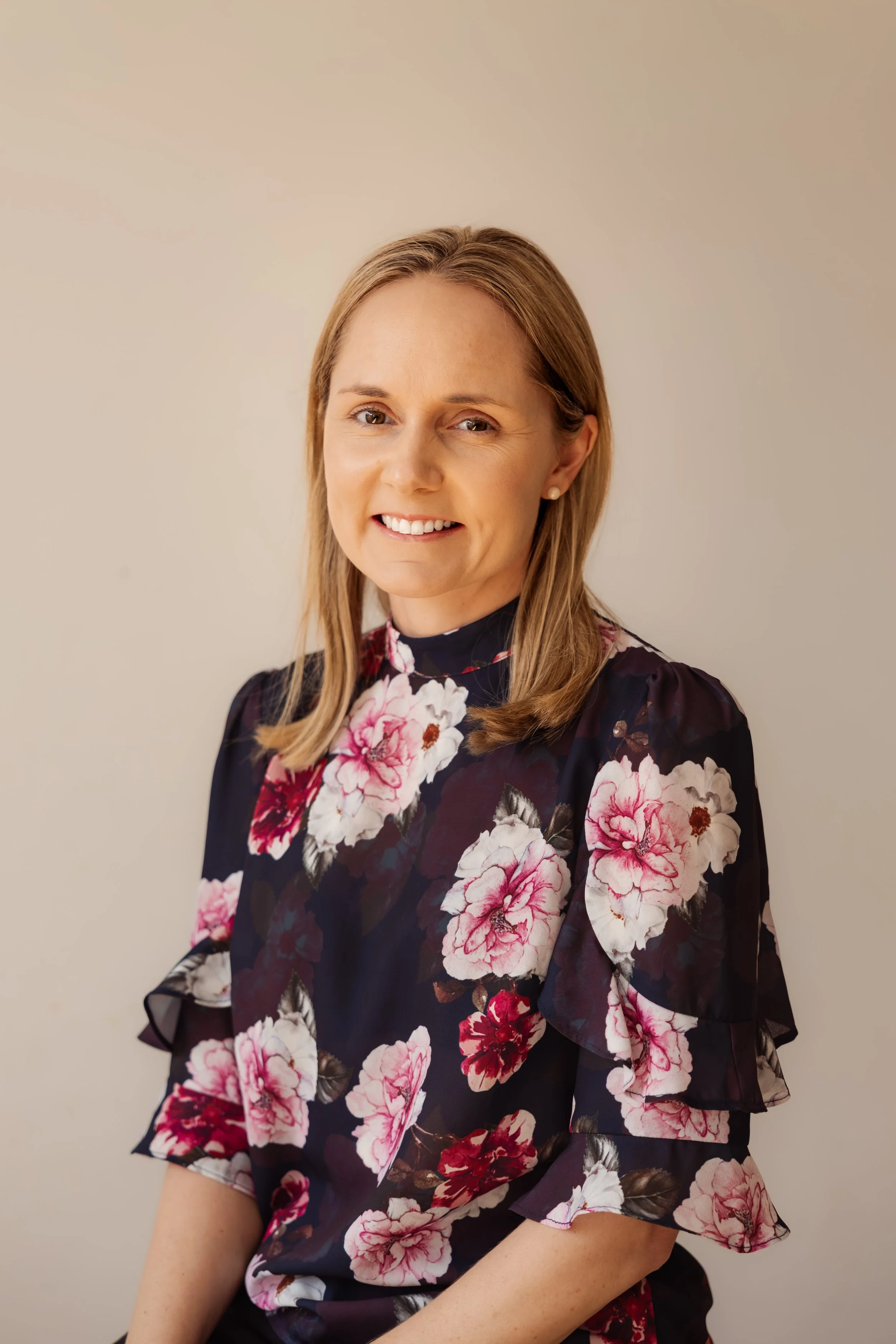 A woman with blonde hair and pearl earrings smiling, wearing a dark floral dress with pink, white, and red flowers, sitting against a plain beige background.