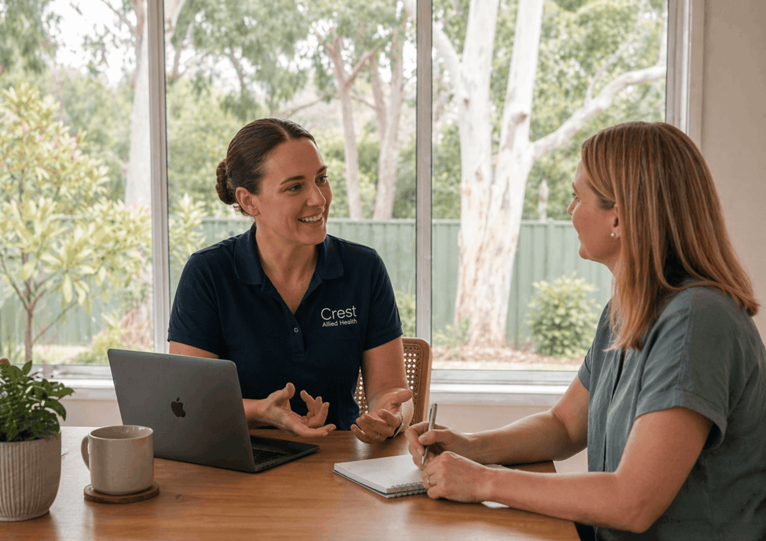 A woman with brown hair in a bun wearing a navy polo shirt with 'Crest Allied Health' logo is talking to another woman with reddish-brown hair, sitting at a wooden table near a window with a garden view. There is a laptop, a notebook, a pen, a mug, and a potted plant on the table.
