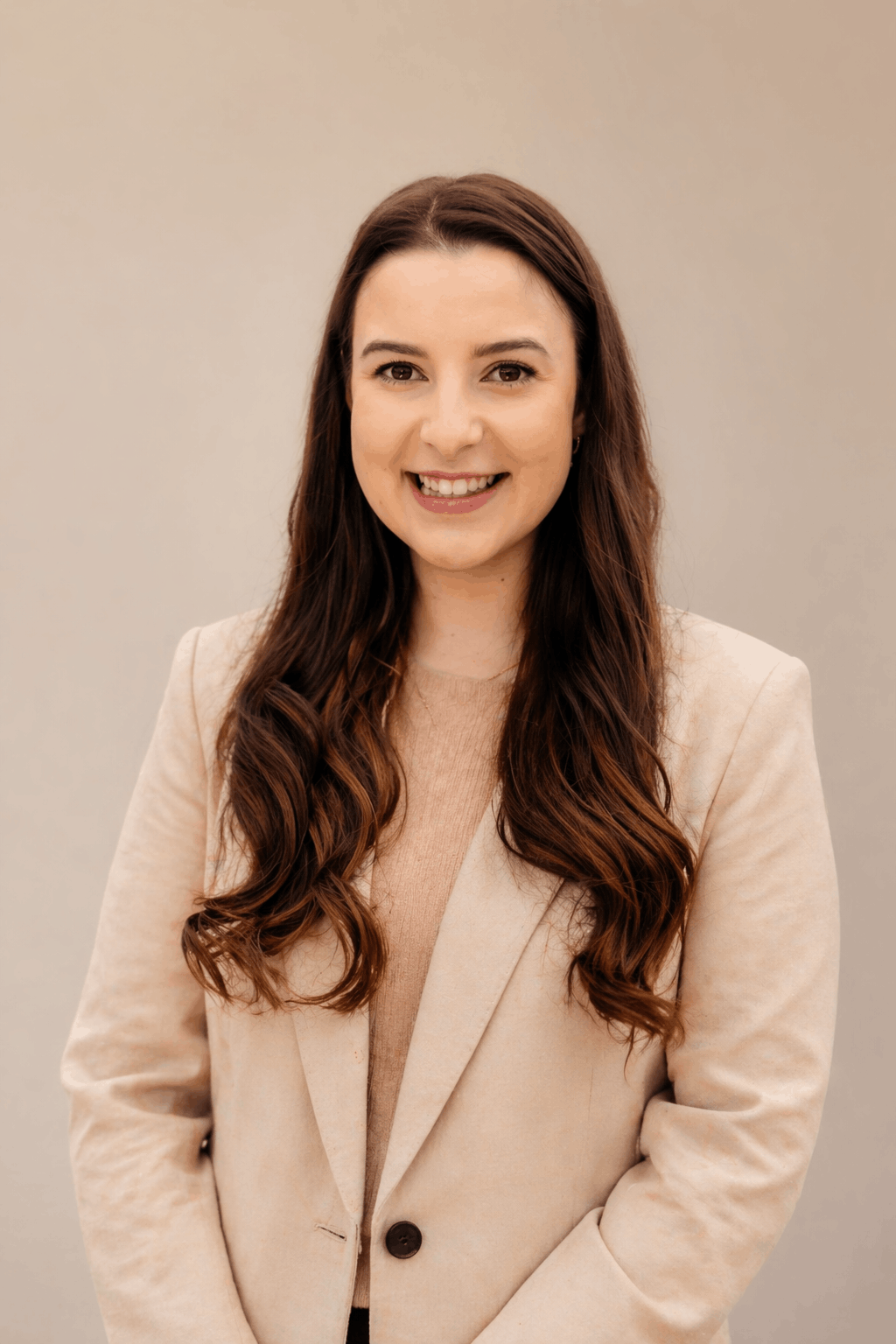 A young woman with long, wavy brown hair smiling and dressed in a beige blazer and top, standing against a neutral background.