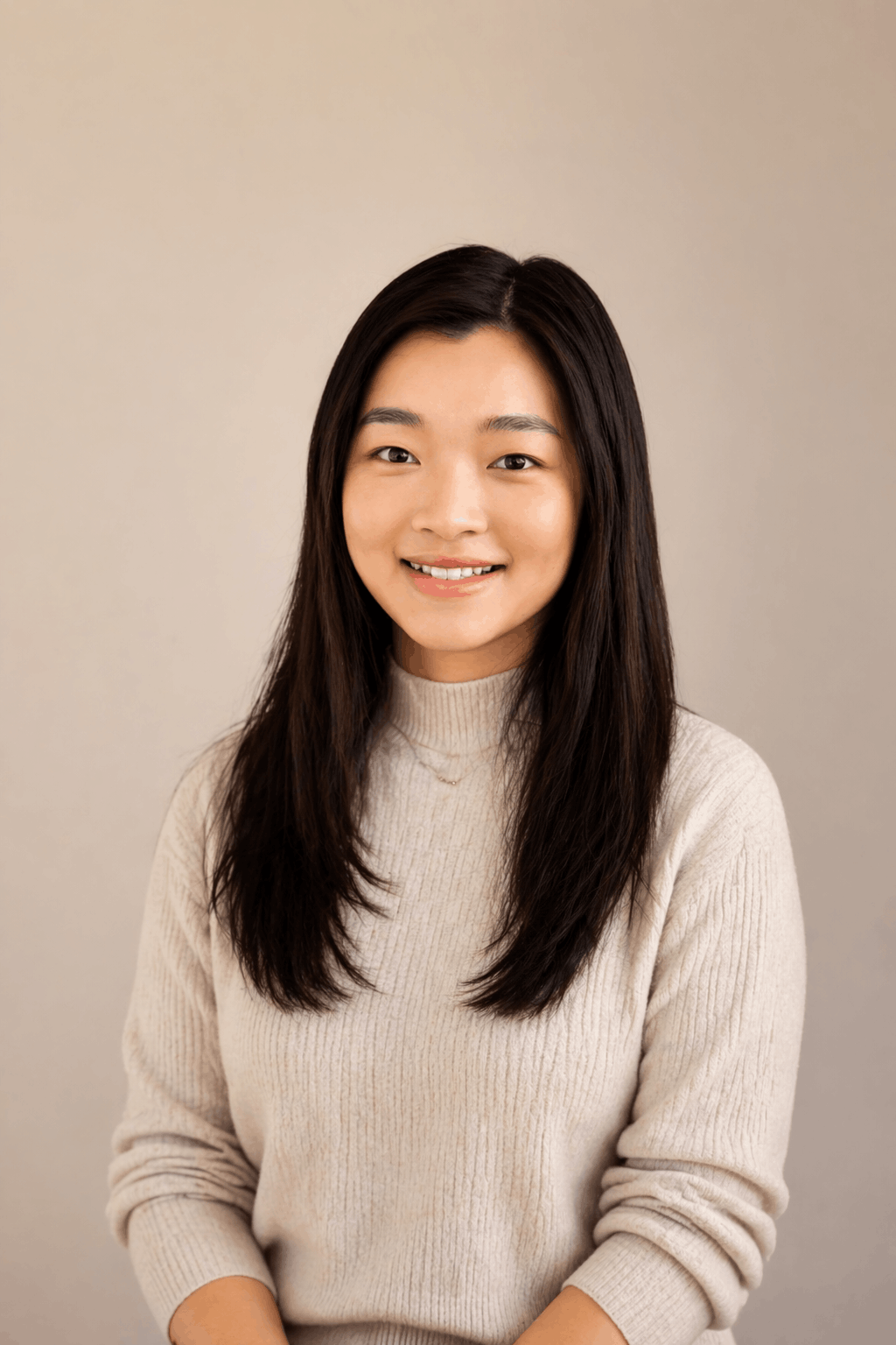 A young woman with long dark hair, smiling, wearing a beige ribbed sweater, standing against a neutral background.