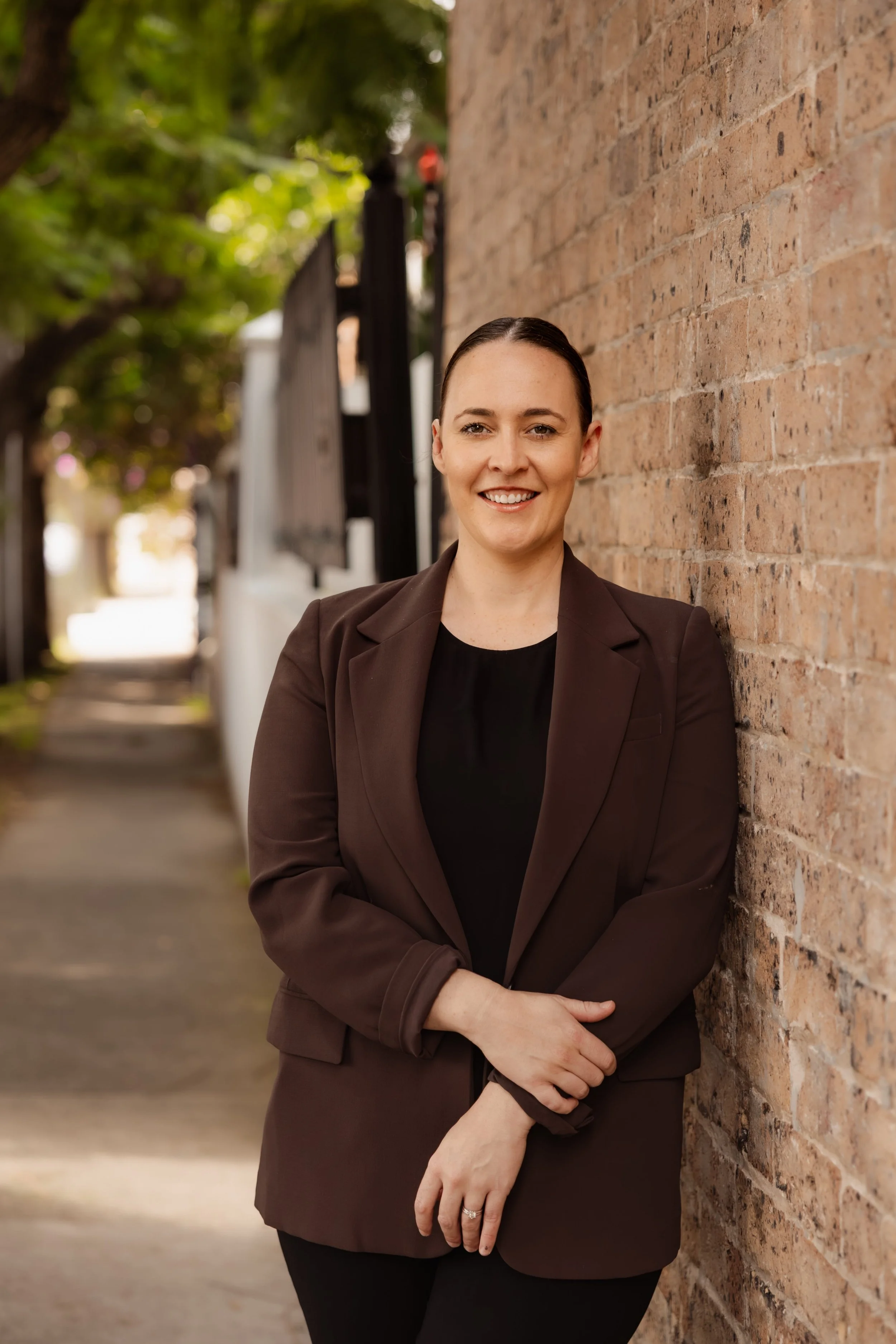 A smiling woman with dark hair in a bun, wearing a brown blazer and black top, standing with arms crossed in front of a brick wall outdoors with trees in the background.