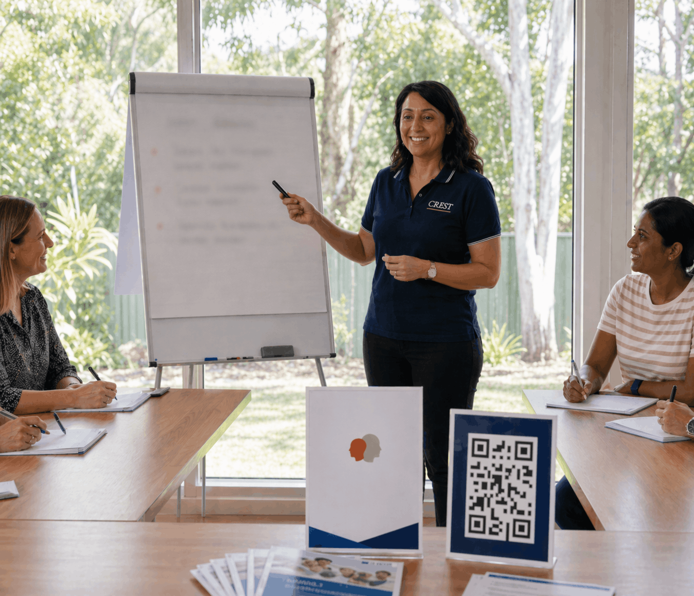 A woman standing at a presentation board, smiling, giving a presentation to three women seated at a conference table with notebooks and pens, in a bright room with large windows showing trees outside.