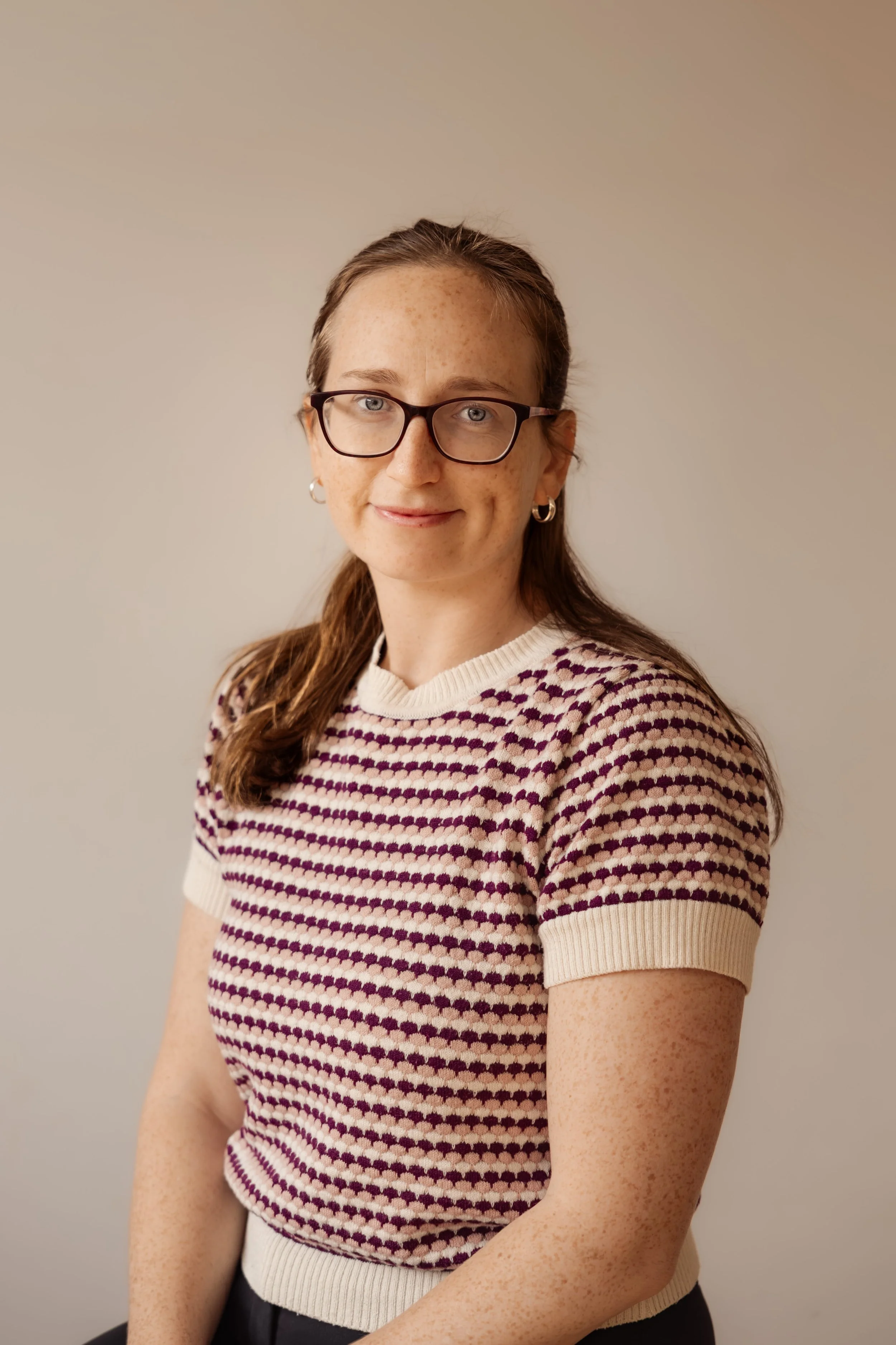 A woman with long brown hair, glasses, and hoop earrings, wearing a short-sleeved, patterned sweater with a cream and dark purple design, standing against a plain beige background.