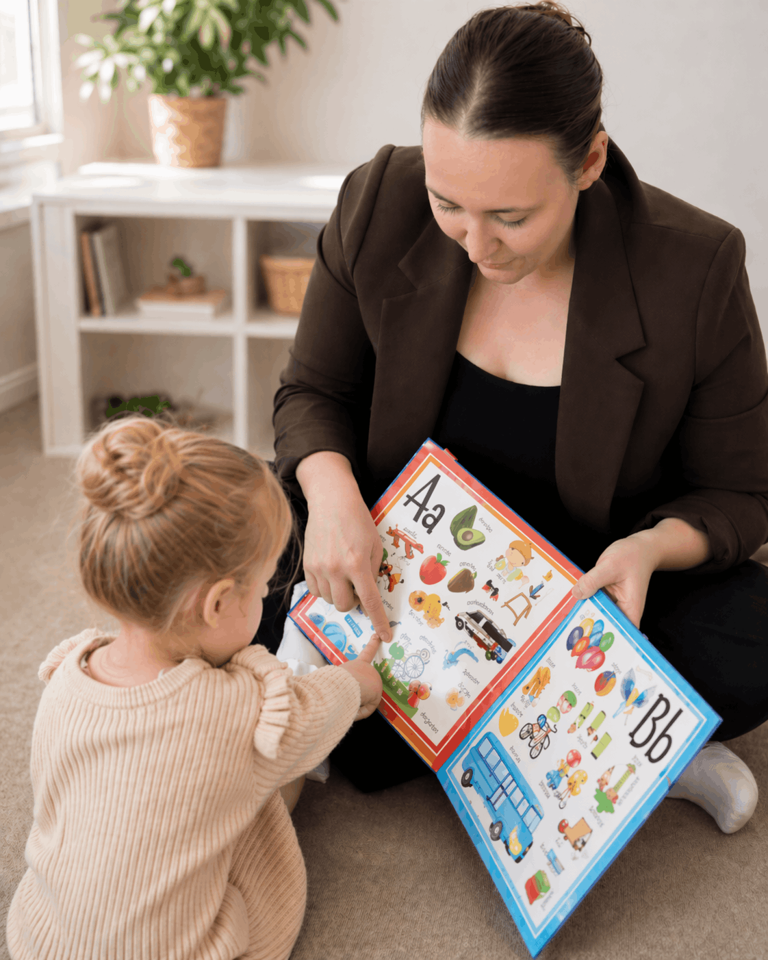 Crest Allied Health _ Sydney A woman and a young girl sitting on the floor, engaged in a learning activity with a colorful alphabet book, pointing at pictures of animals, objects, and vehicles.