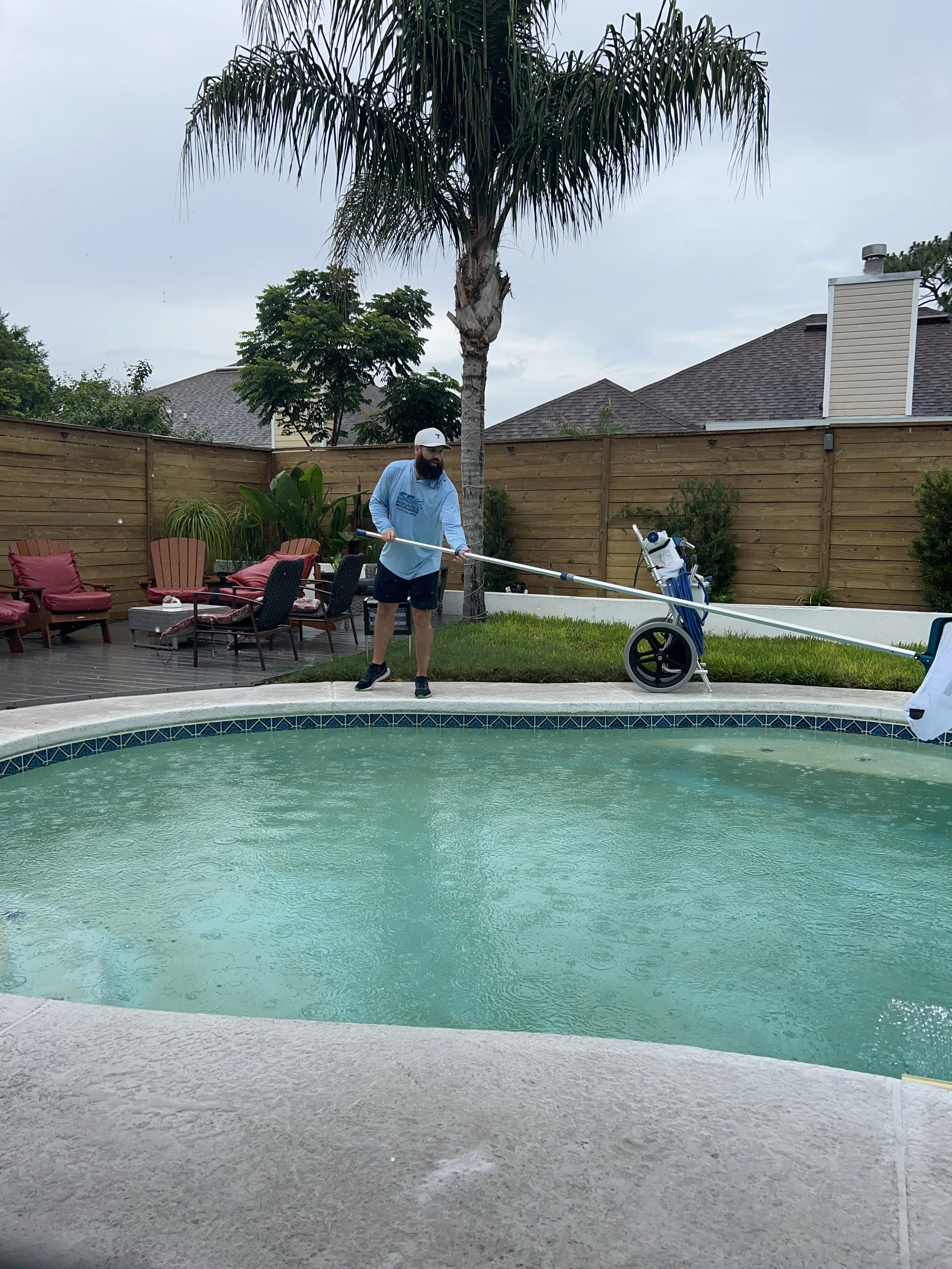 A man with a beard wearing a white cap, blue long sleeve shirt, and black shorts is cleaning a backyard swimming pool with a long pool cleaning pole. The pool has turquoise water and a white concrete deck. In the background, there is a wooden fence, some outdoor furniture with pink cushions, tropical plants including a tall palm tree, and a neighborhood with houses and a cloudy sky.