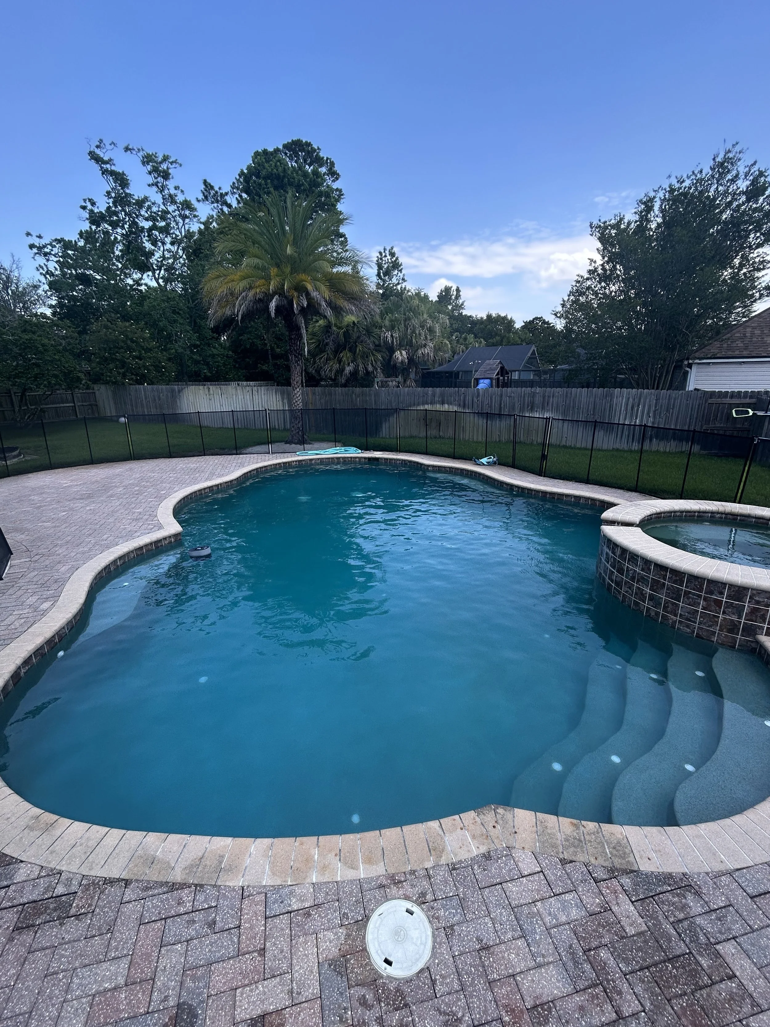 Backyard view with swimming pool, hot tub, brick patio, surrounded by a wooden fence, with trees and houses in the background, under a blue sky with some clouds.