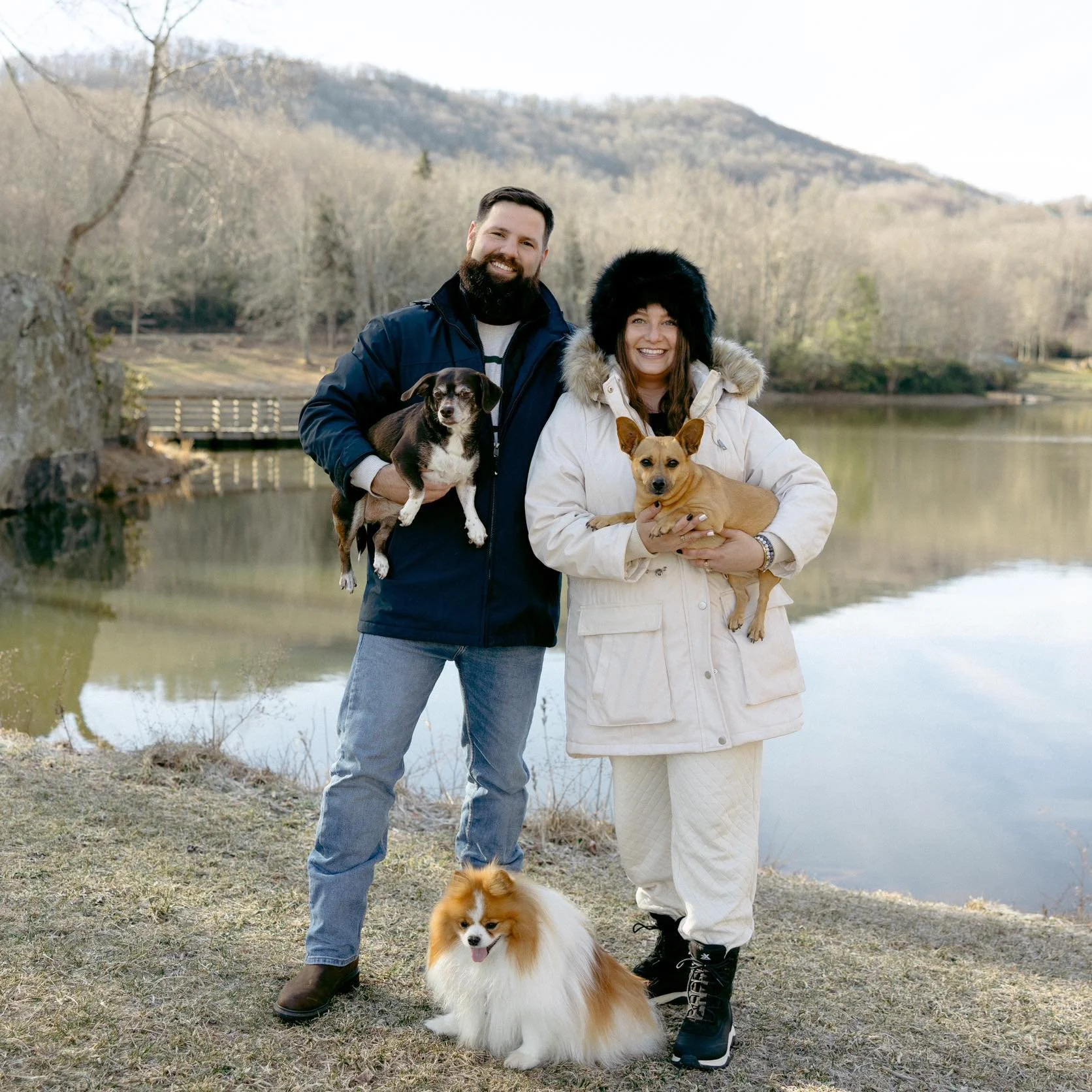 A smiling man and woman stand outdoors near a river, each holding a dog. The man has dark hair and a beard, wearing a dark blue jacket and jeans, holding a small black and white dog. The woman has brown hair, wearing a white coat and black boots, holding a light brown dog. A fluffy Pomeranian is sitting on the ground in front of them. In the background, there are trees, a wooden bridge, and hills under a cloudy sky.