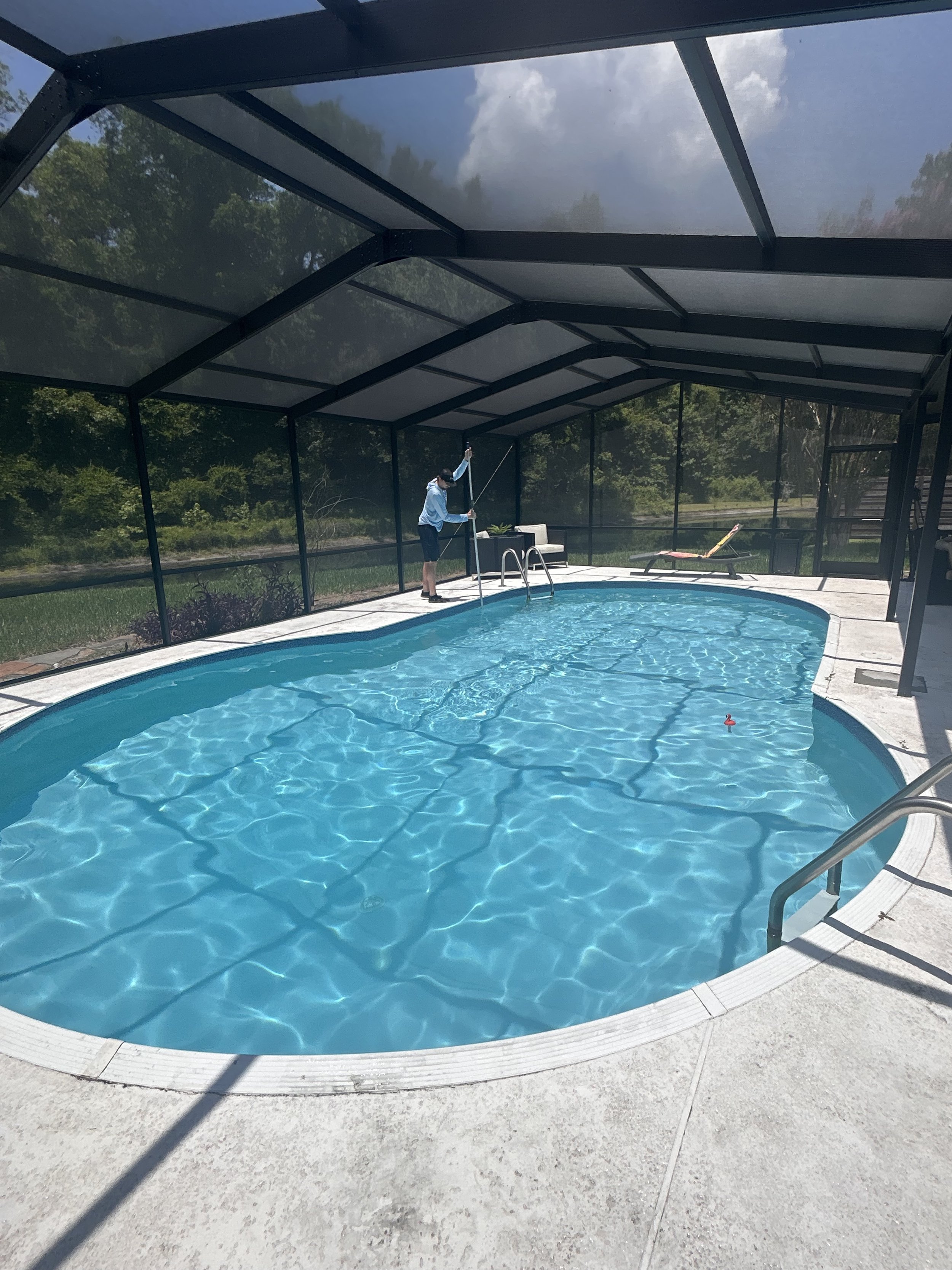 A man cleaning a backyard swimming pool under a screened enclosure on a sunny day.
