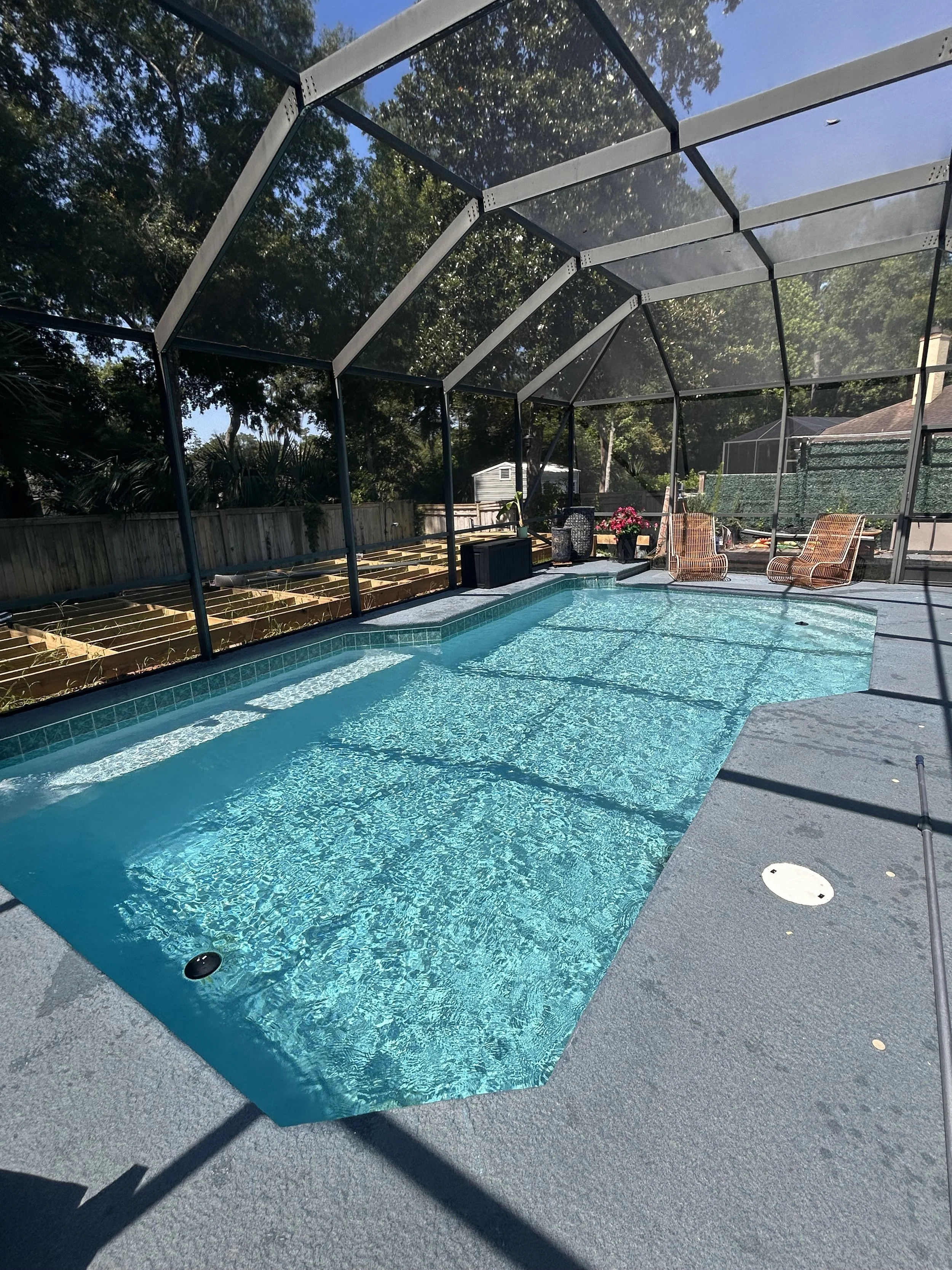 A backyard swimming pool enclosed by a screened structure with a seating area including two wicker chairs, a small table with a flower pot, and various potted plants.
