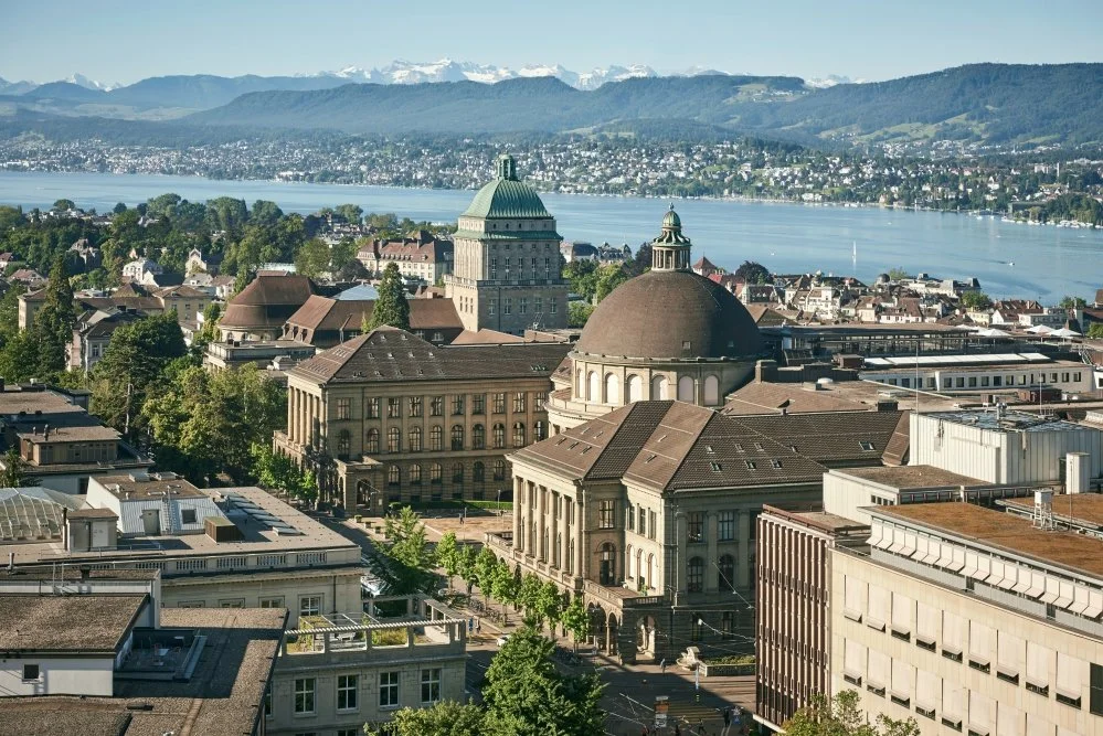 A cityscape with historic buildings, a river, and mountains in the background under a clear sky.