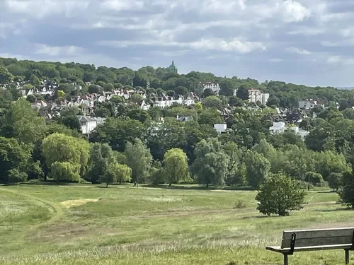 A scenic view of a city on a hill with trees and houses, over a grassy park with a bench in the foreground, under a cloudy sky.