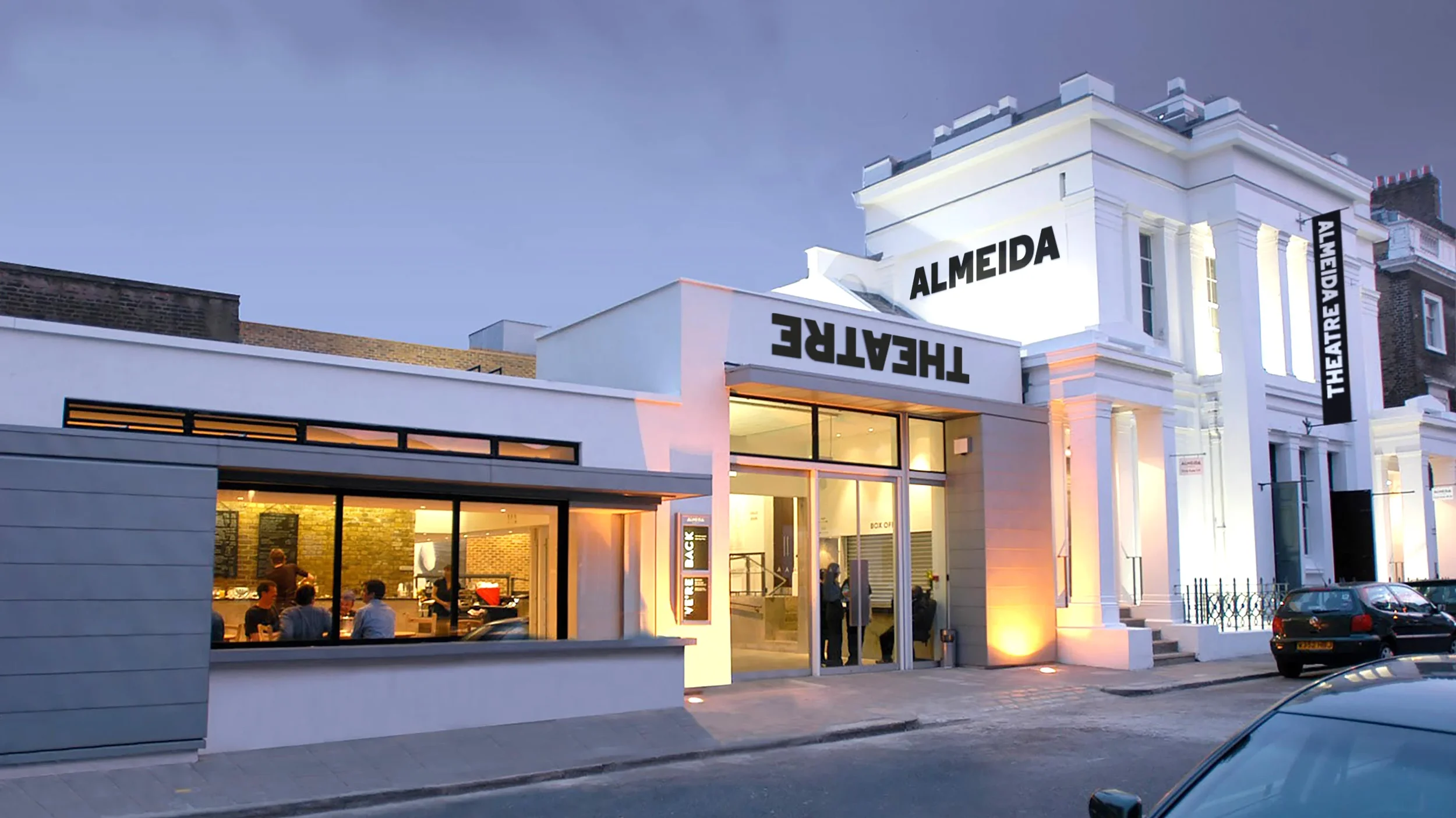 Exterior of Almeida Theatre building at dusk with illuminated signage, large glass windows revealing people inside, and a small parking area with cars in front.