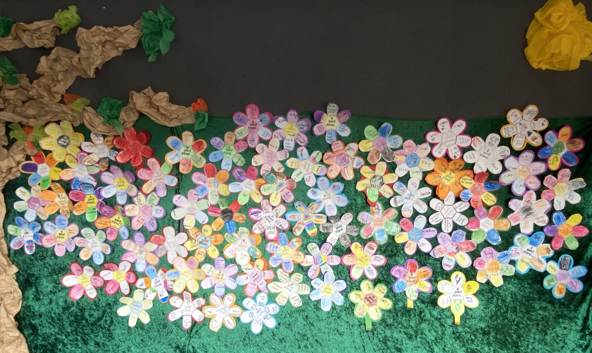Colorful paper flowers with handwritten notes and messages, decorated on a green background, with crumpled paper to simulate clouds or mountains at the top left corner.
